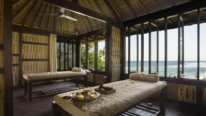 A room with a wooden floor, a bed with a patterned bedspread, a bench, and a table with a tray of flowers and a bowl. - Jeeva Klui Resort (Senggigi)