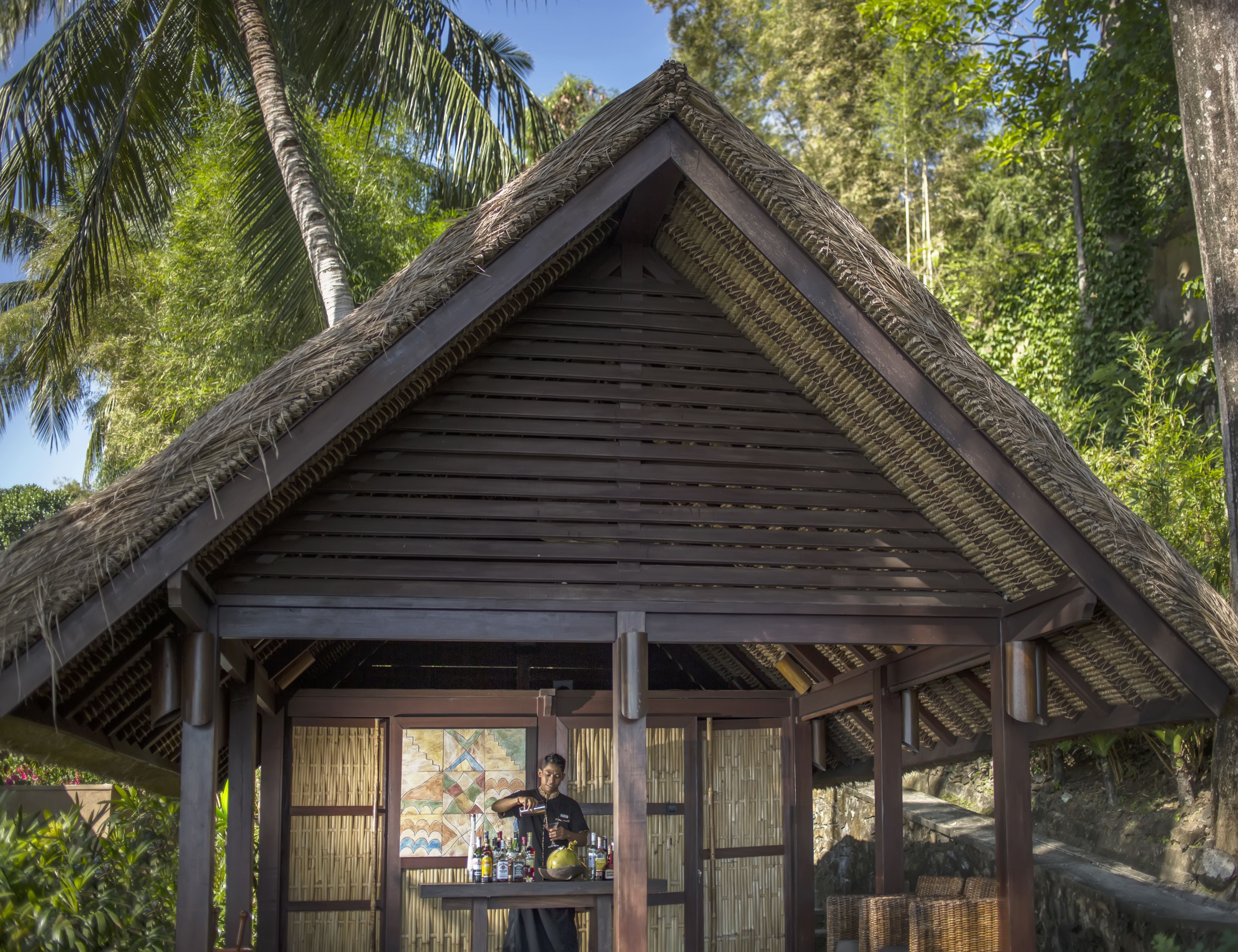 A wooden structure with a thatched roof, a bar area with bottles, and wicker furniture.