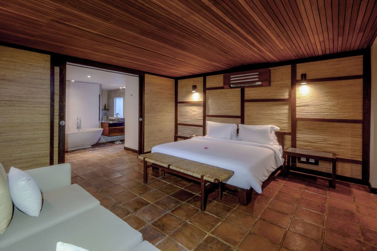 A bedroom with a wooden ceiling, a large bed, a bench, and a small bedside table.
