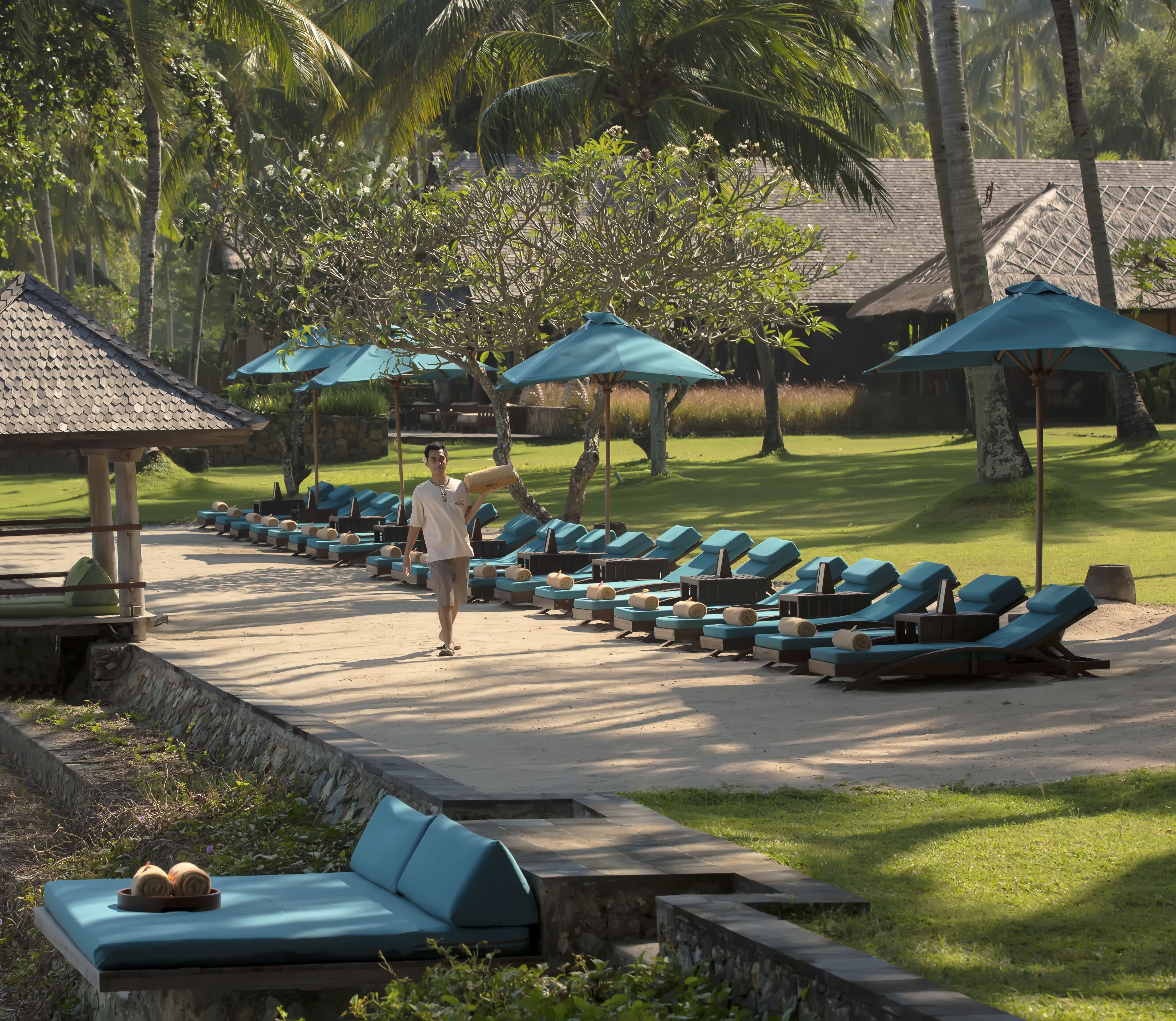 Un resort con filas de sillas de playa bajo sombrillas azules, un área de piscina y abundante vegetación.