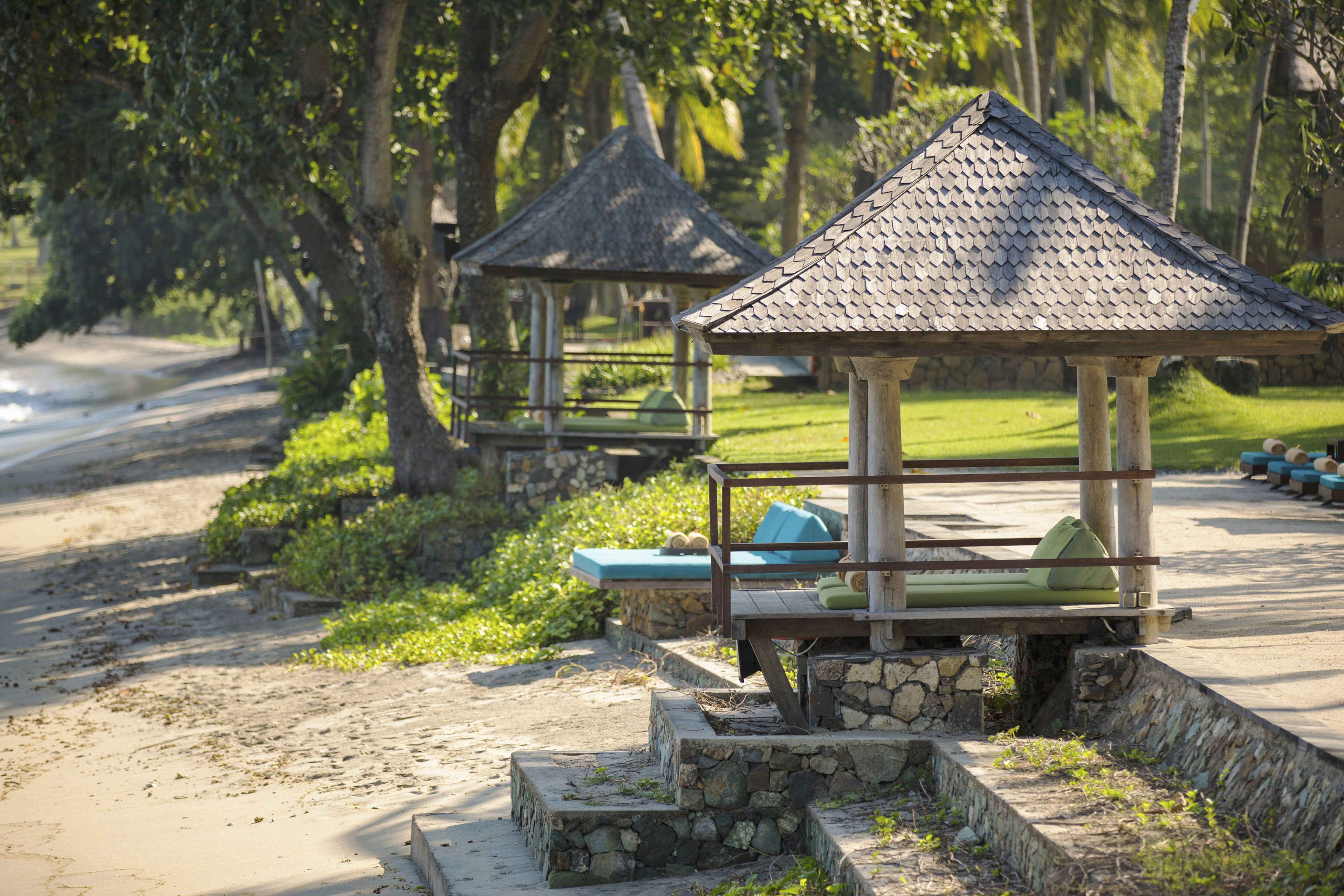 A beachside resort with stone steps leading to a gazebo with a thatched roof, surrounded by lush greenery and a sandy path.