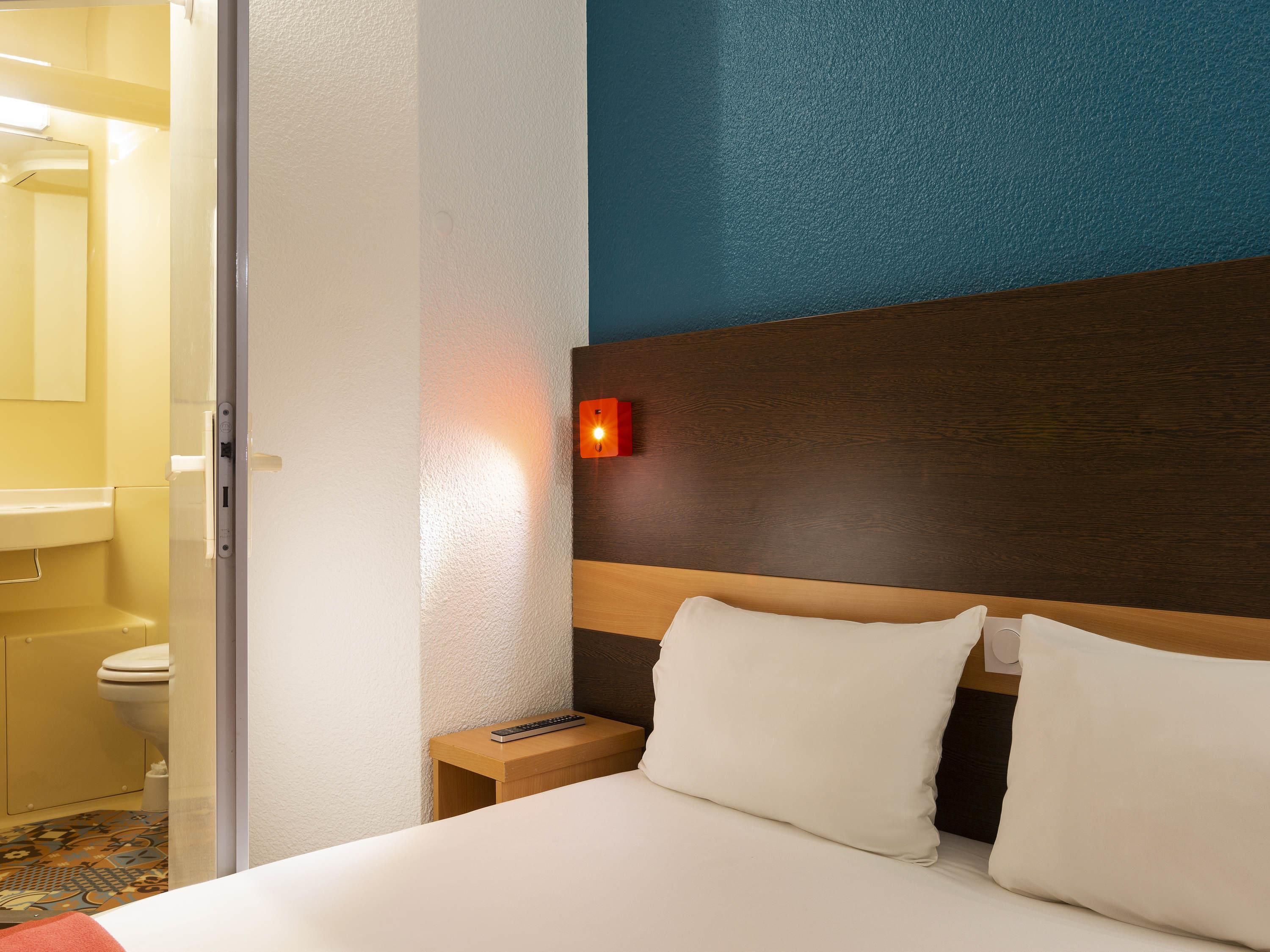 A hotel room with a bed, a nightstand, a wall-mounted light, and a bathroom visible through an open door.
