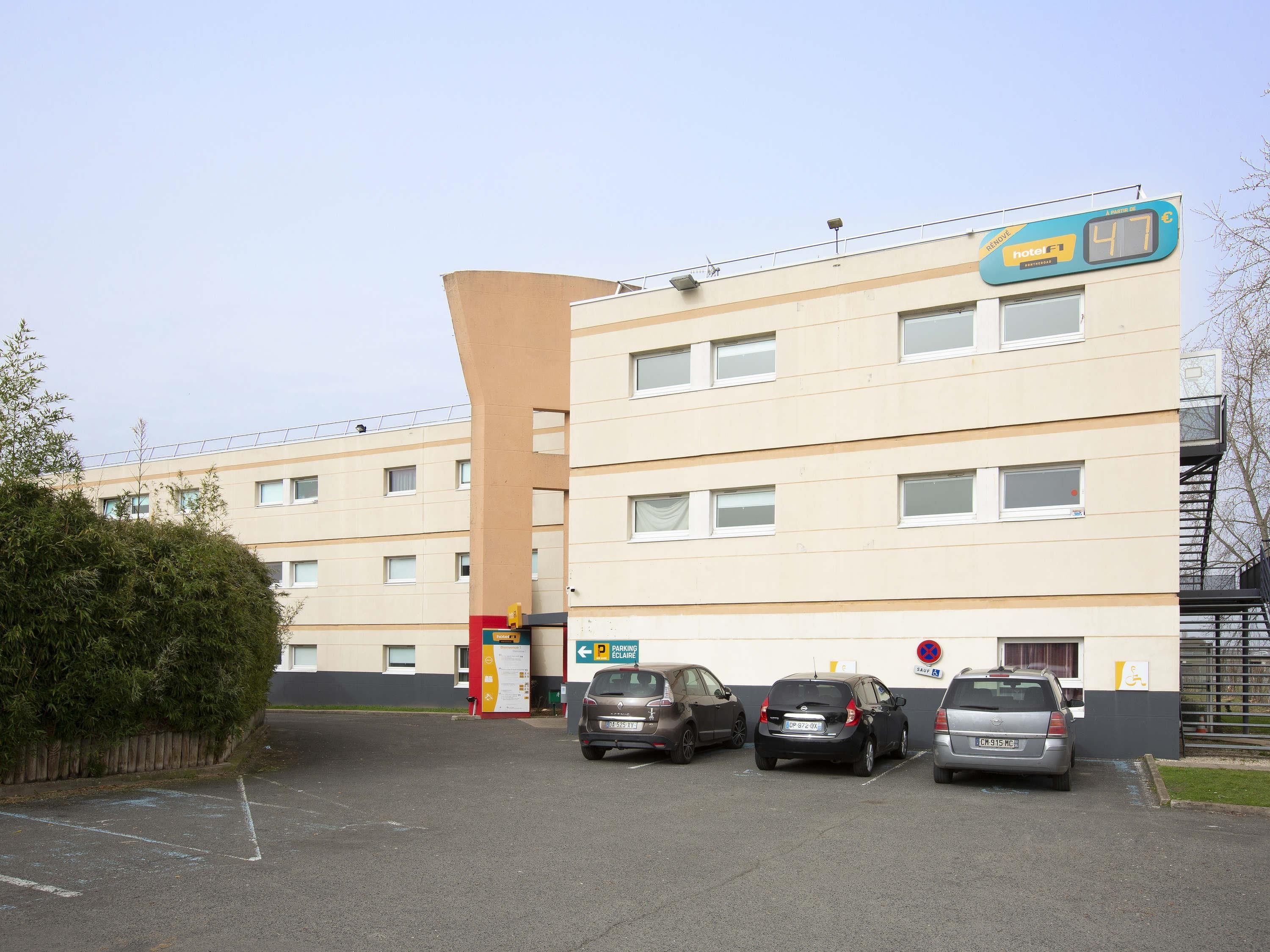 Un hotel de varios pisos con diseño moderno, un estacionamiento con varios autos y un letrero que indica un precio de 47 euros.