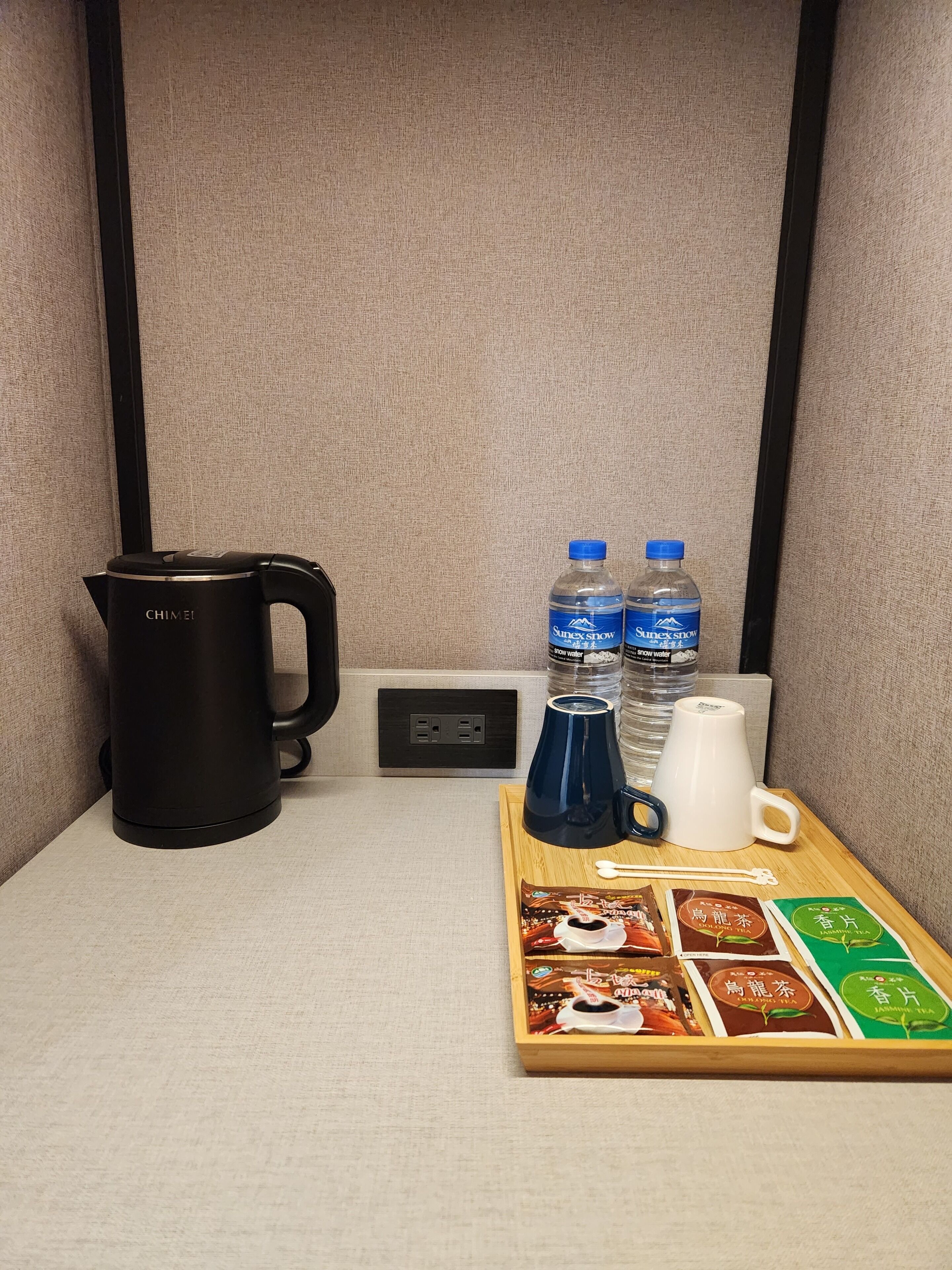 Una cuina americana amb una olla negra Chiemel, dues ampolles d'aigua Suprikol, una cafetera blava, una tassa blanca i un safata amb sobres de te.