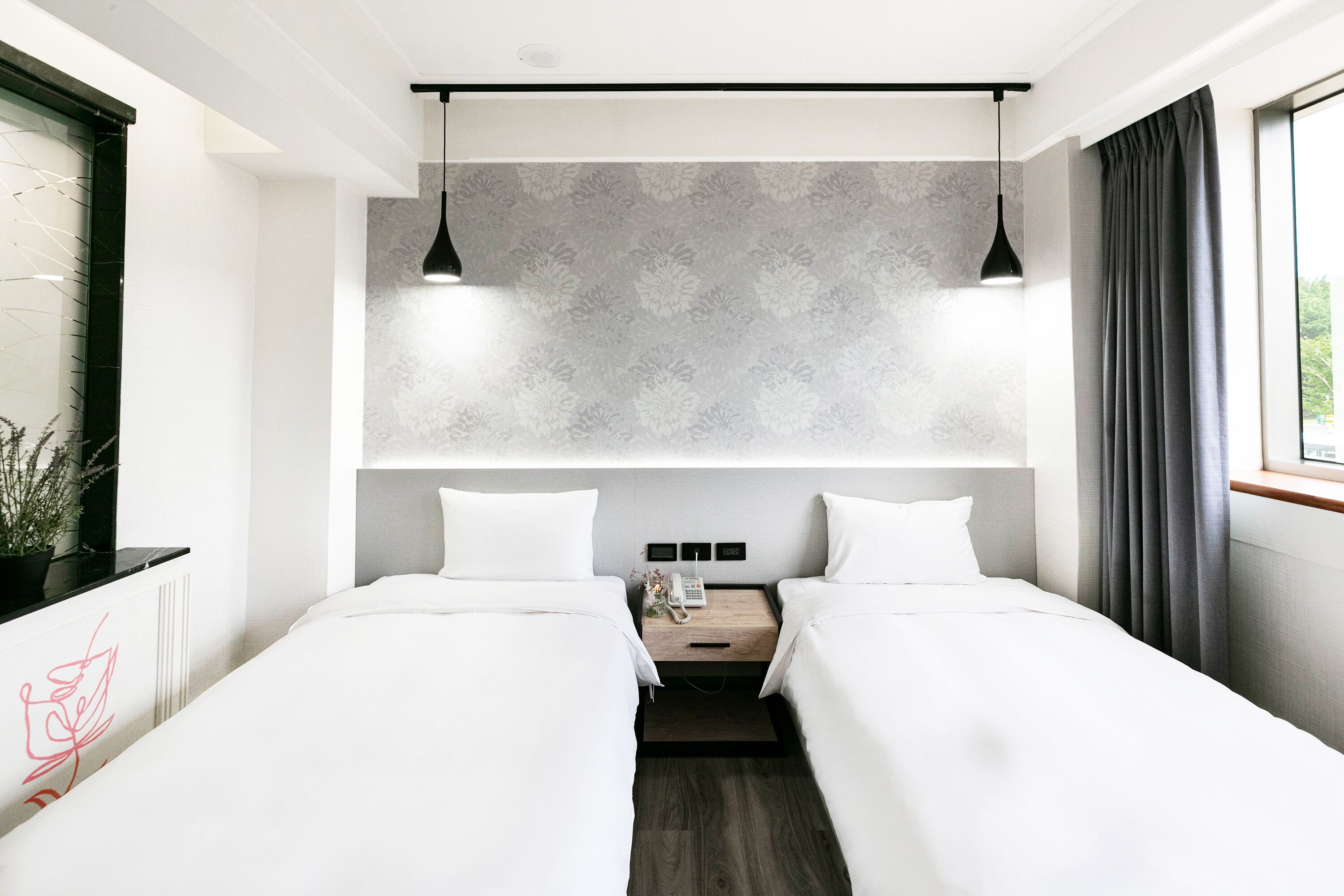 Ein modernes Hotelzimmer mit zwei Einzelbetten, einem kleinen Nachttischchen mit Telefon, einer Wandleuchte und einem Fenster mit Vorhängen.