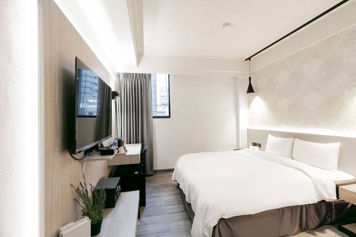 A modern hotel room with a large bed, a desk with a flat-screen TV, a window with curtains, and a headboard with built-in lighting.