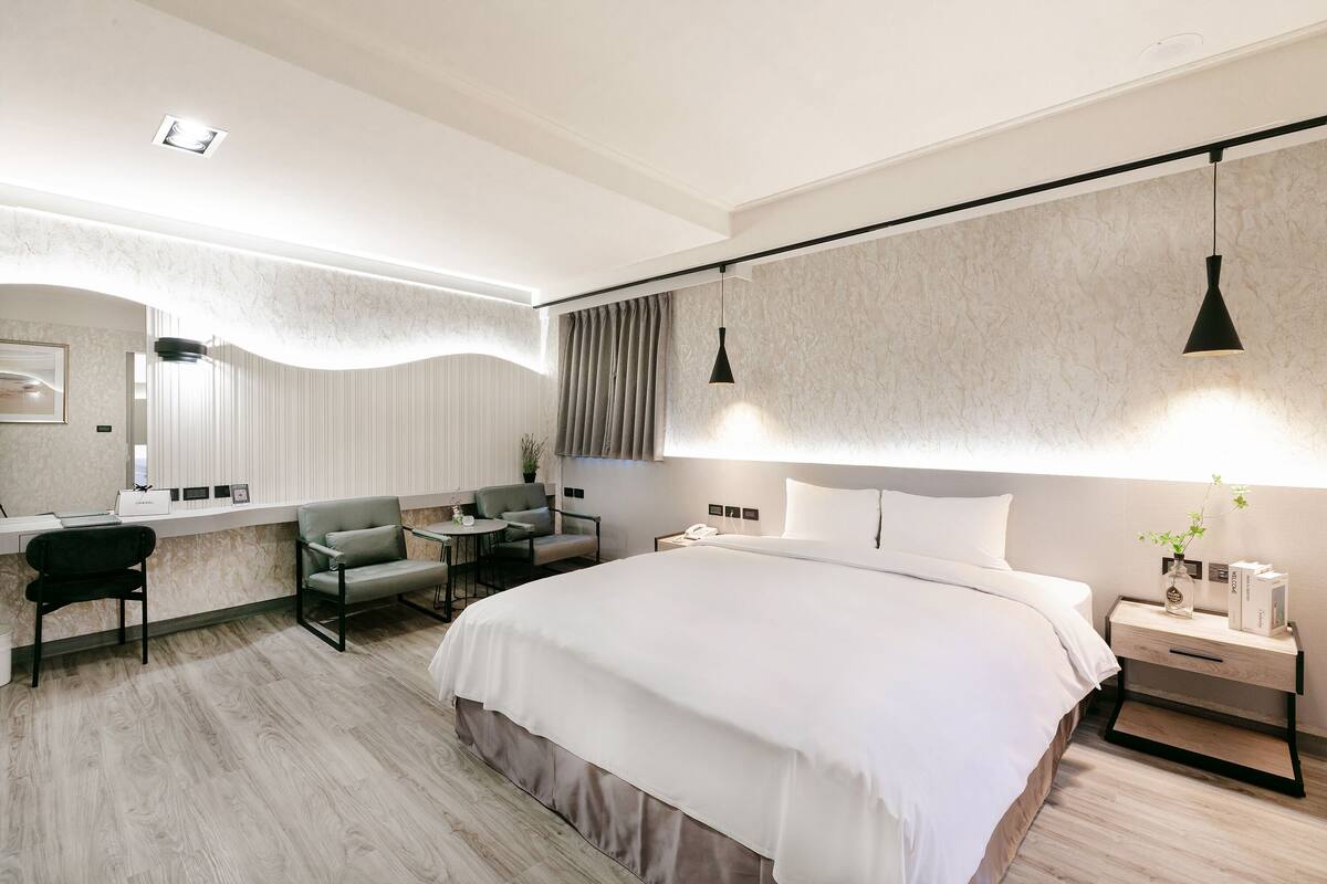 A modern hotel room with a large bed, a desk with a chair, a small table, and a nightstand with a lamp.