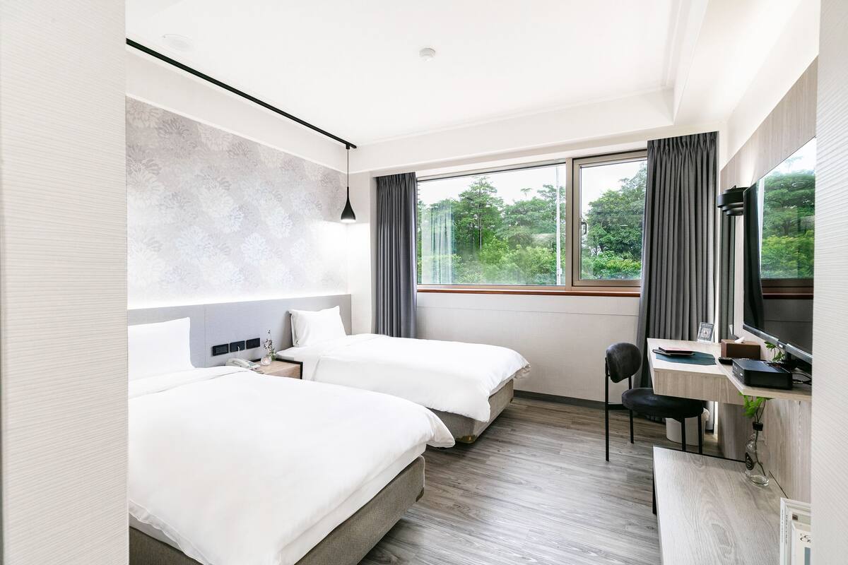 A modern hotel room with two beds, a desk, and a large window with curtains.