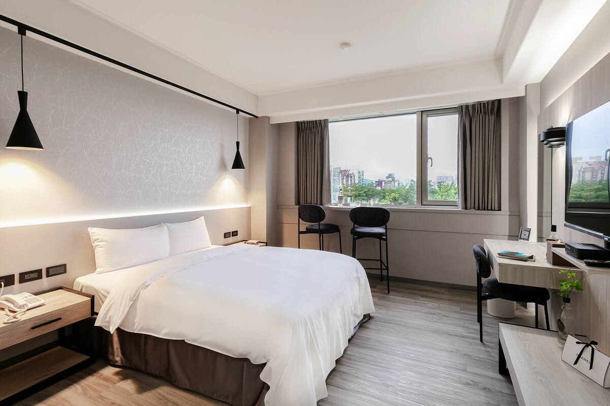 A hotel room with a bed, bedside table, desk, chair, and a large window with a city view.