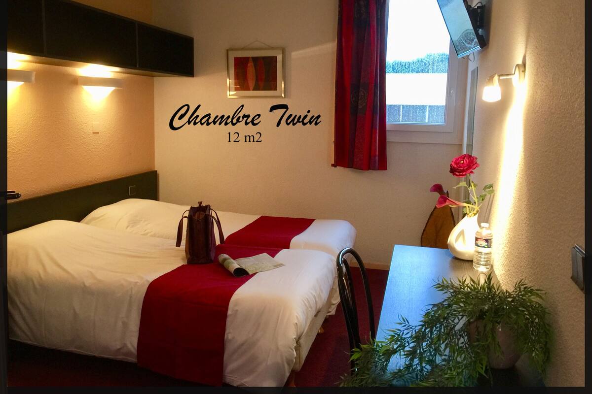 A twin room with a bed, a desk, a TV, a window with curtains, and a wall-mounted sign indicating the room size.