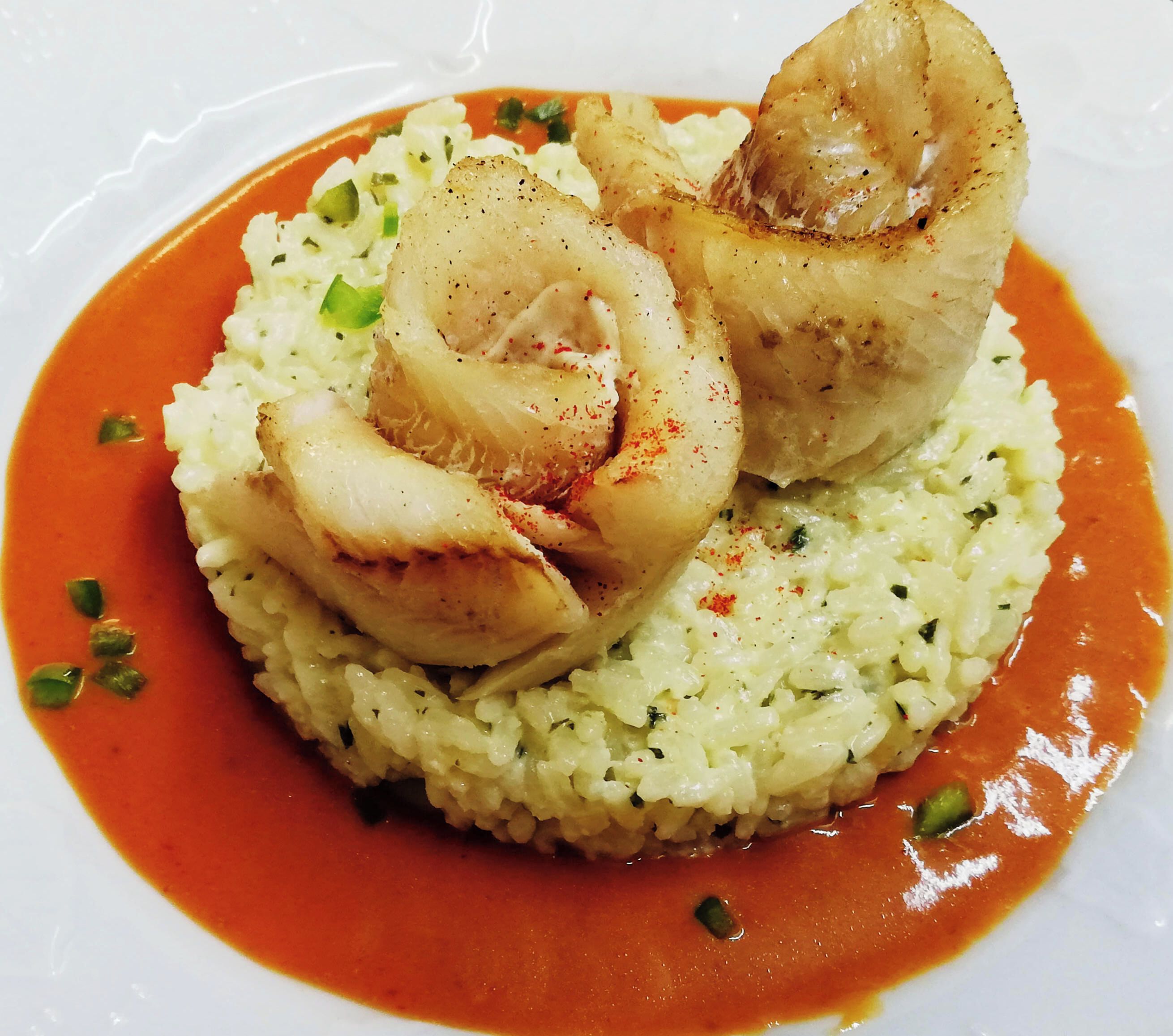 Un plato con base blanca, cubierto por una sustancia cremosa y salsa roja, decorado con trozos verdes.