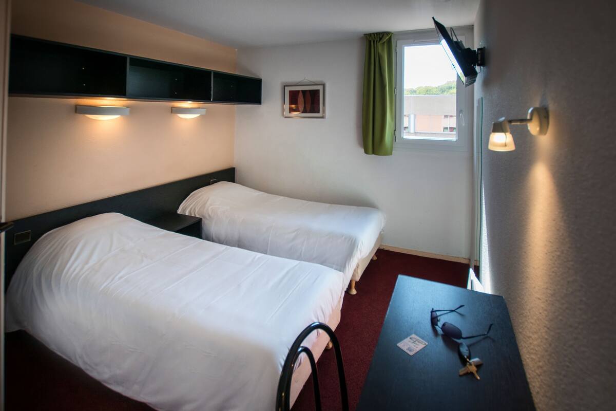 A hotel room with two beds, a small table, a pair of sunglasses, and a window with a curtain.
