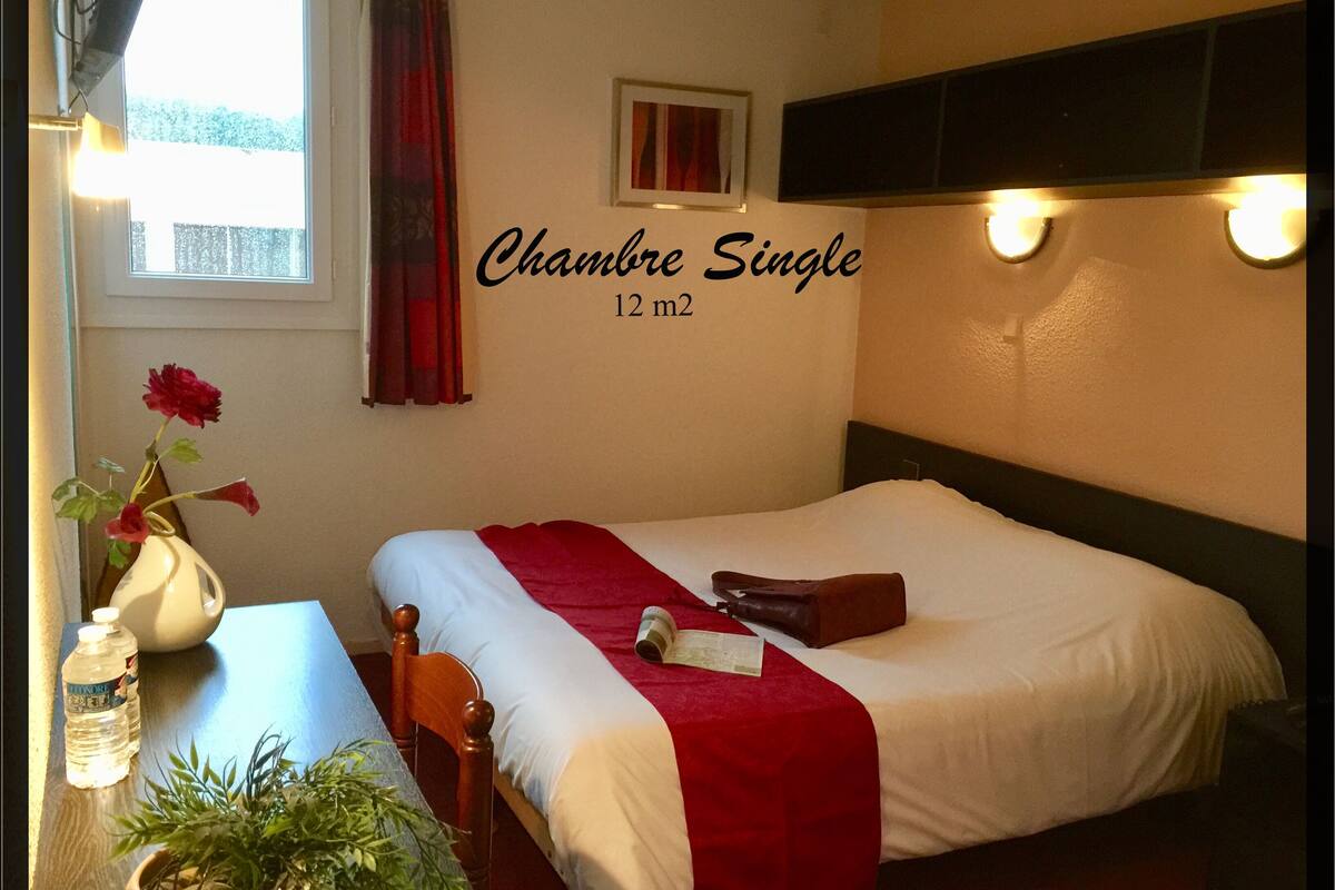 A single room with a bed, a bedside table with a vase of flowers, and a window with curtains.