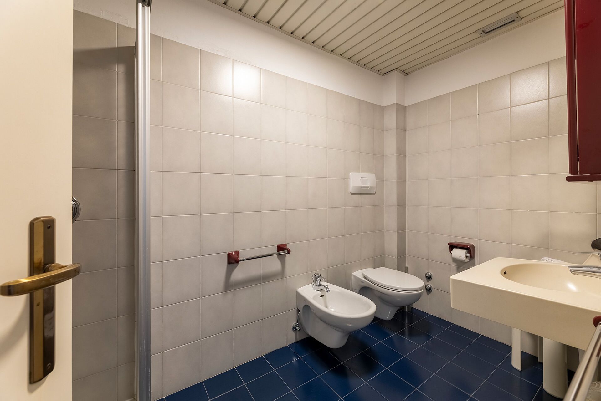 A small, modern bathroom with white tiles, a blue floor, a bidet, a toilet, and a sink.