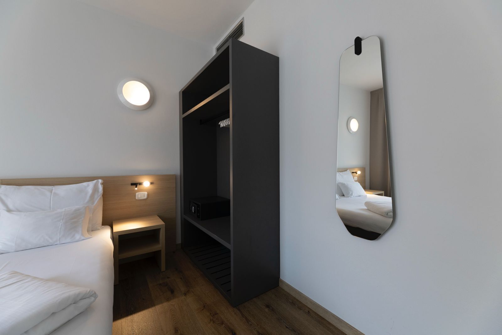 A modern hotel room with a bed, bedside table, built-in wardrobe, and a wall-mounted mirror.