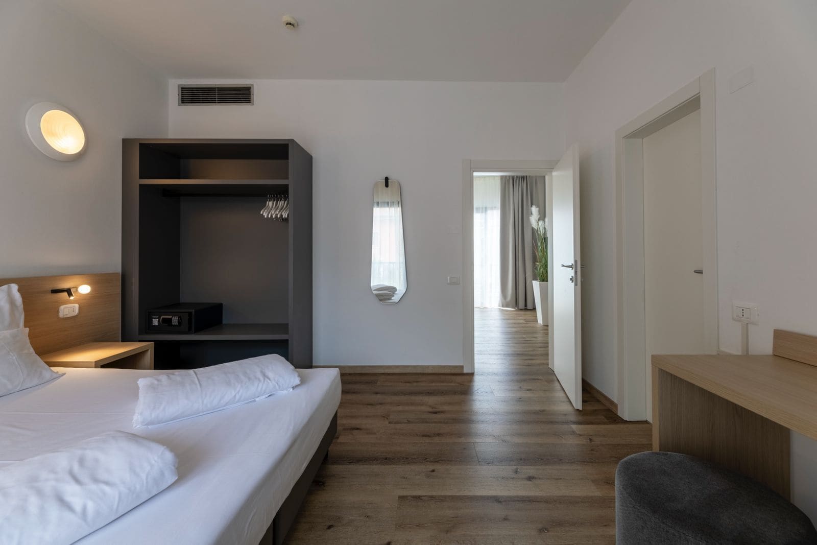 A modern hotel room with a bed, bedside table, built-in wardrobe, and a view of the corridor.