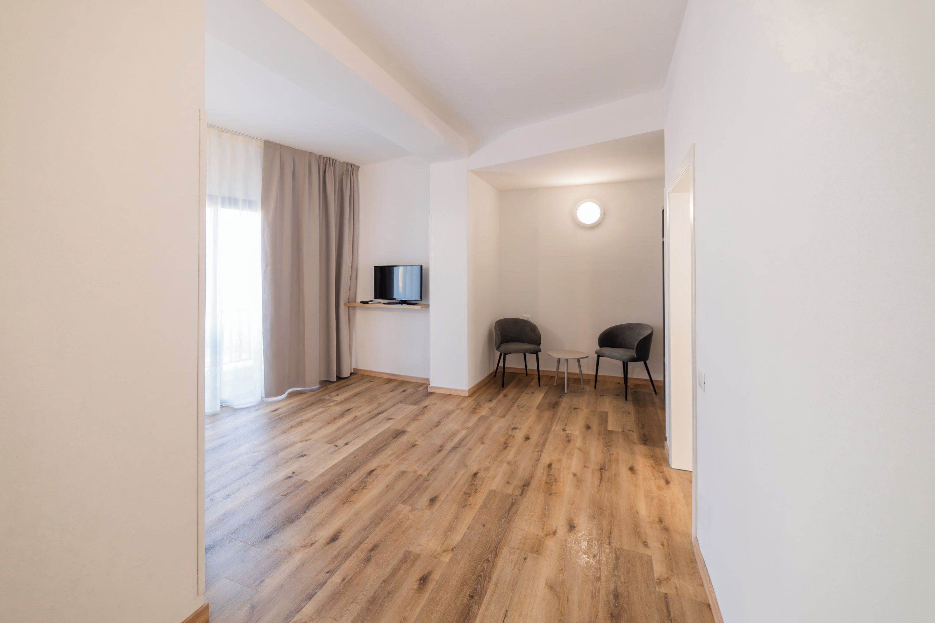 A room with wooden flooring, a television on a stand, two chairs, and a round light fixture on the wall.