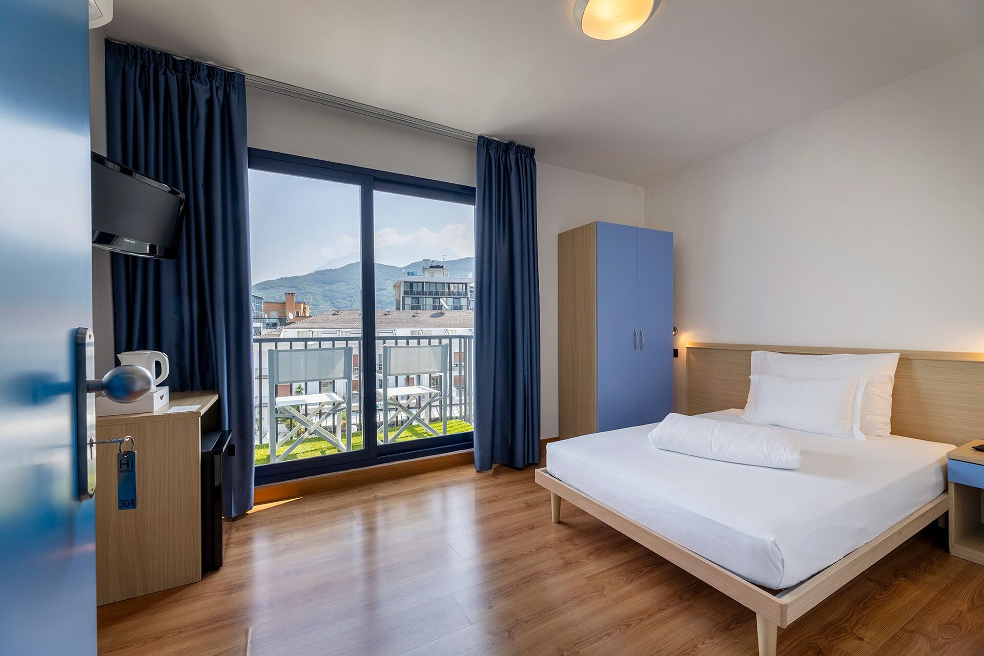 Ein Hotelzimmer mit Bett, Schreibtisch mit Kaffeemaschine, Kleiderschrank und großem Fenster mit Vorhängen.