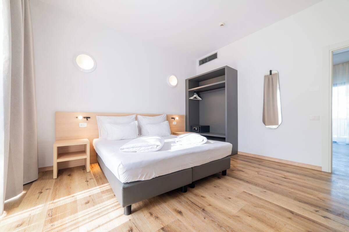 A modern hotel room with a large bed, a bedside table, a wardrobe, and a mirror on the wall.