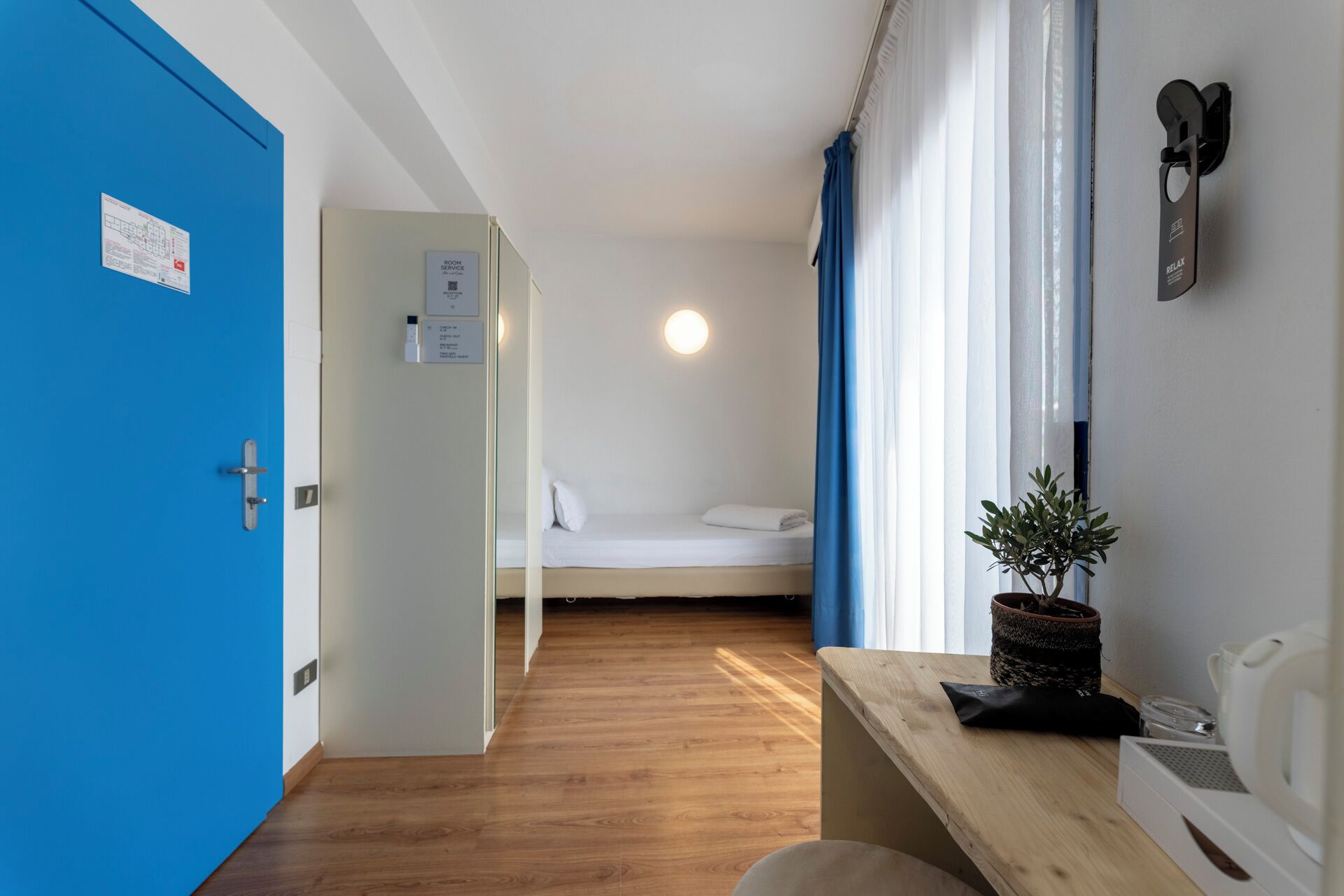 Habitación de hotel con piso de madera, cama con ropa blanca, puerta azul, una pequeña mesa con una planta y una lámpara fijada a la pared.