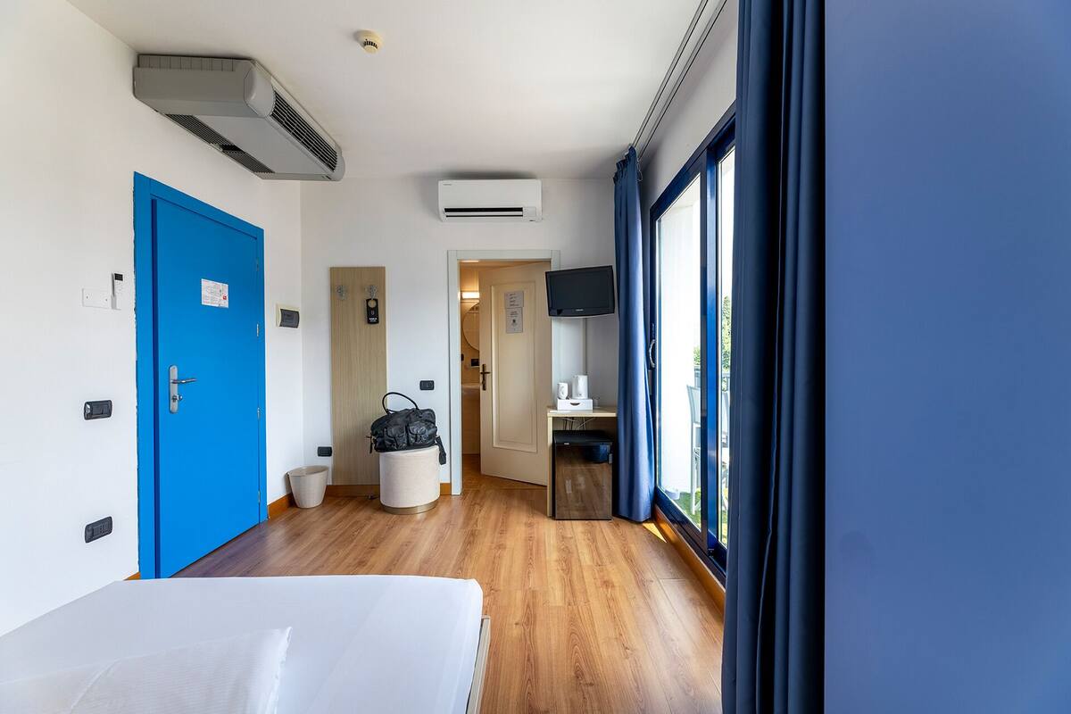 A hotel room with a wooden floor, a blue door, an air conditioning unit, a television, and a window with curtains.