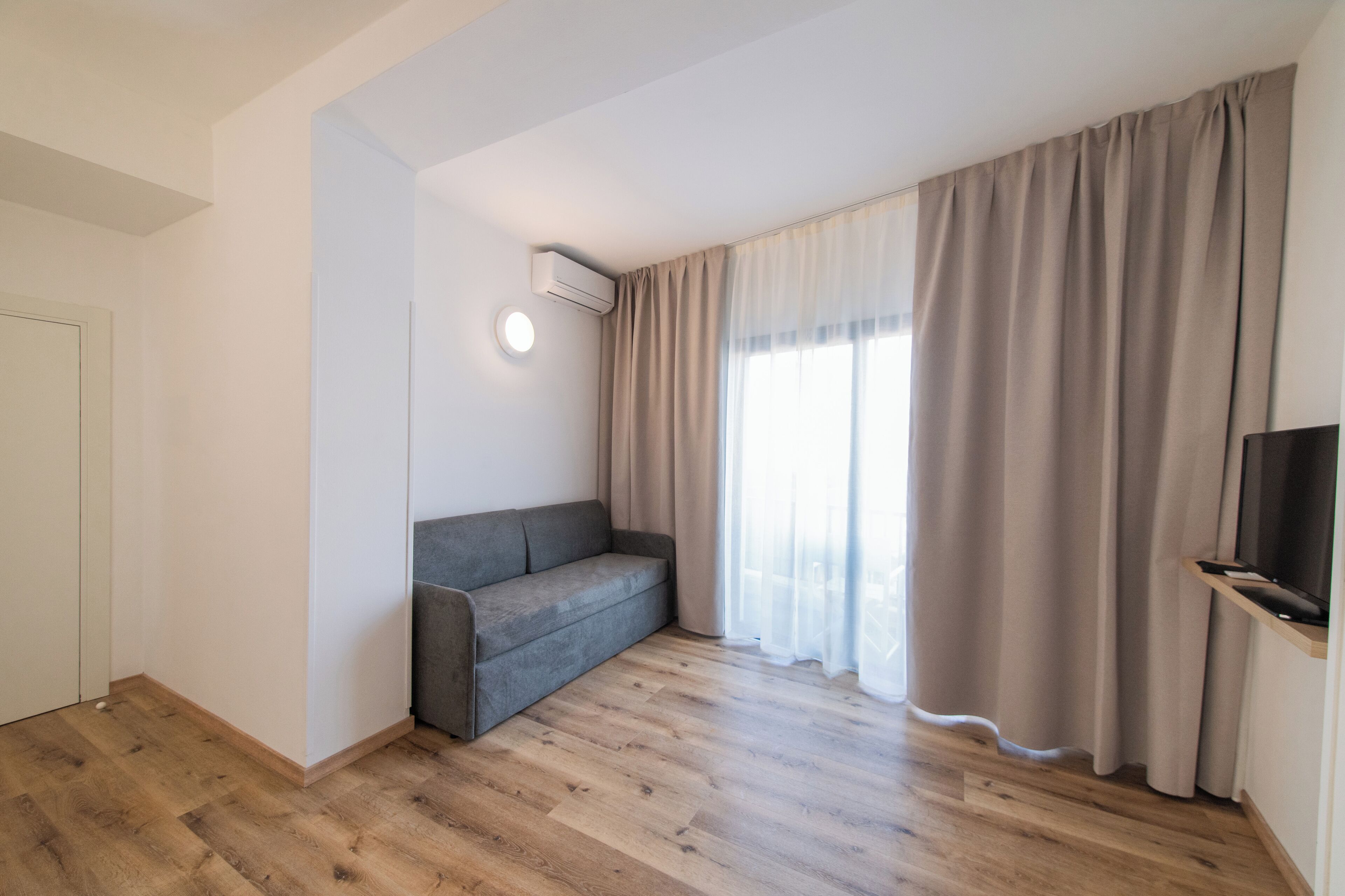 A modern living room with a grey sofa, wooden flooring, a flat-screen TV, and large windows with beige curtains.