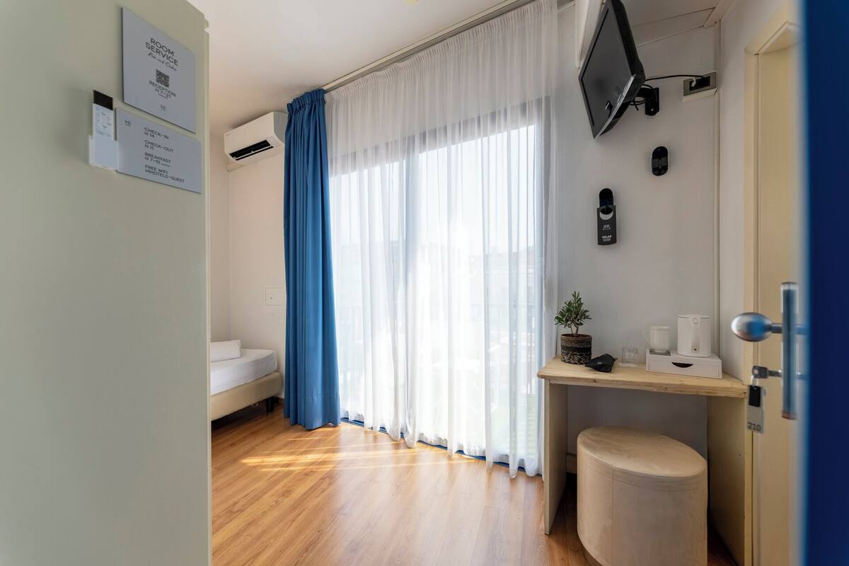 A hotel room with a bed, a desk with a plant, a coffee machine, and a TV mounted on the wall.