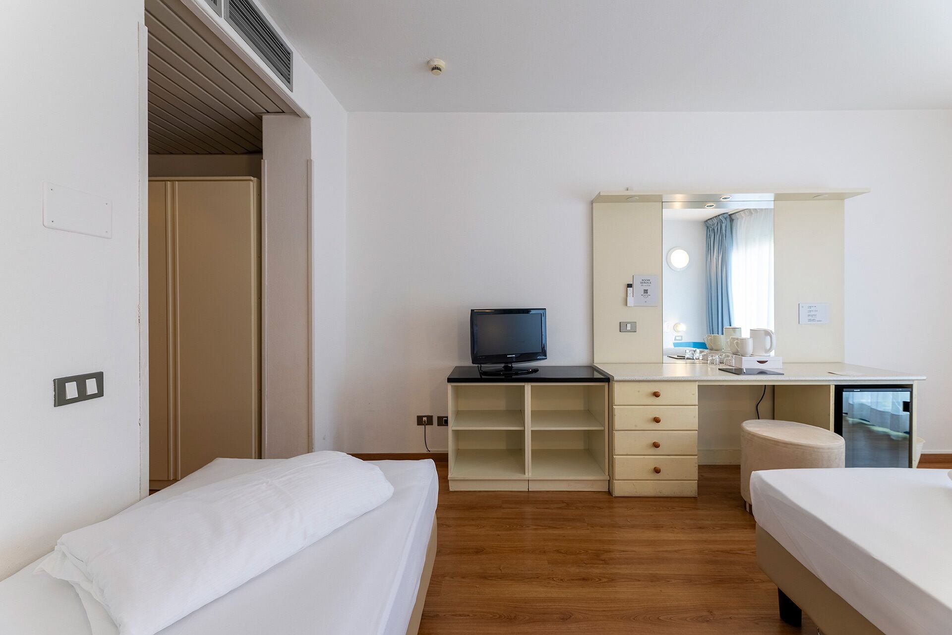 A hotel room with two beds, a desk with a television, a mirror, and a window with curtains.