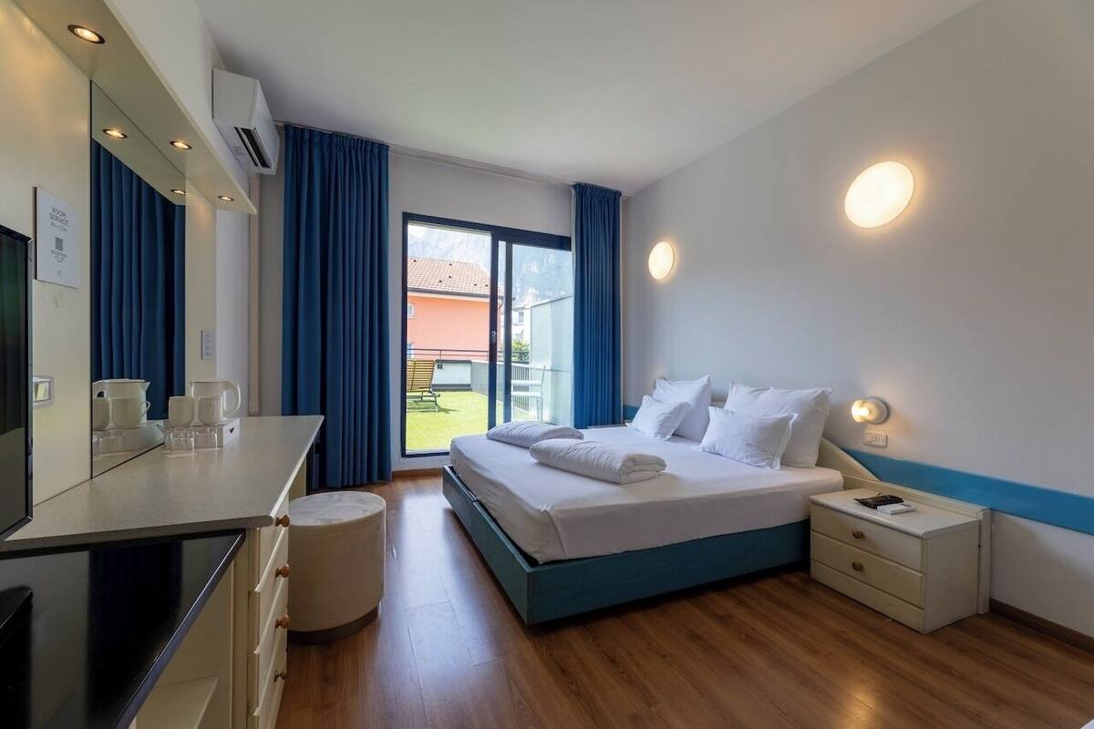 A modern hotel room with a bed, bedside table, and a small stool. There is a window with curtains, a view of the outside, and a flat-screen TV on the left side.