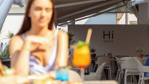 Eine Frau sitzt an einem Tisch mit einem Getränk vor sich, im Hintergrund ein Schild mit der Aufschrift 'Hi HOTELS' und eine Reihe von Tischen und Stühlen.