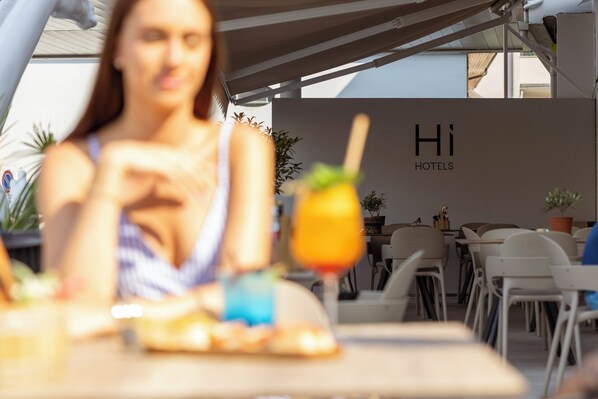 A woman sitting at a table with a drink in front of her, a sign reading "Hi HOTELS" in the background, and a row of chairs and tables.
