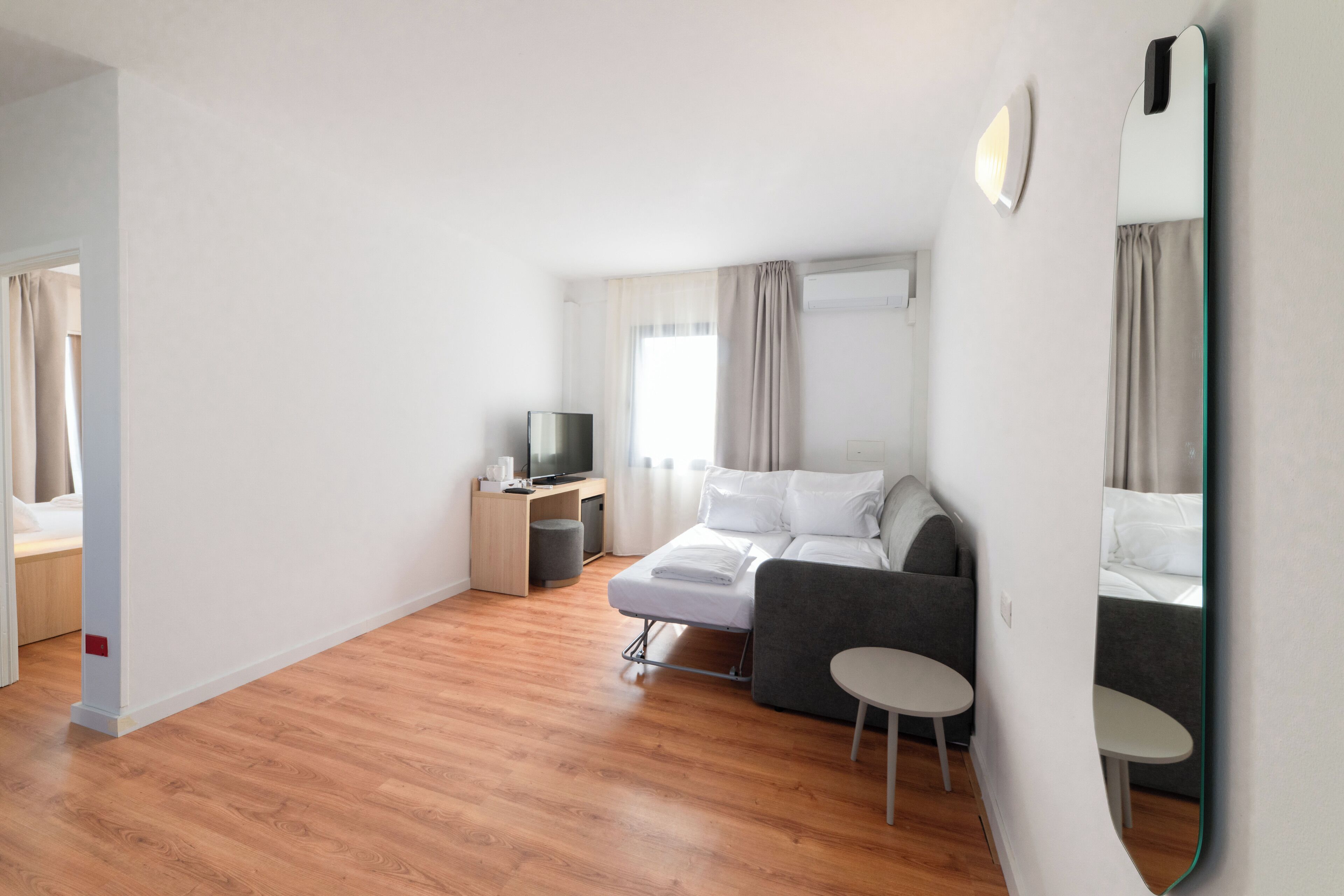A modern hotel room with a wooden floor, a bed with white linens, a grey sofa, a small table, a TV, and a mirror.