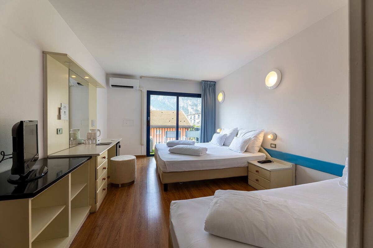 A hotel room with a bed, bedside tables, a desk with a computer, a mirror, and a window with a view of buildings.