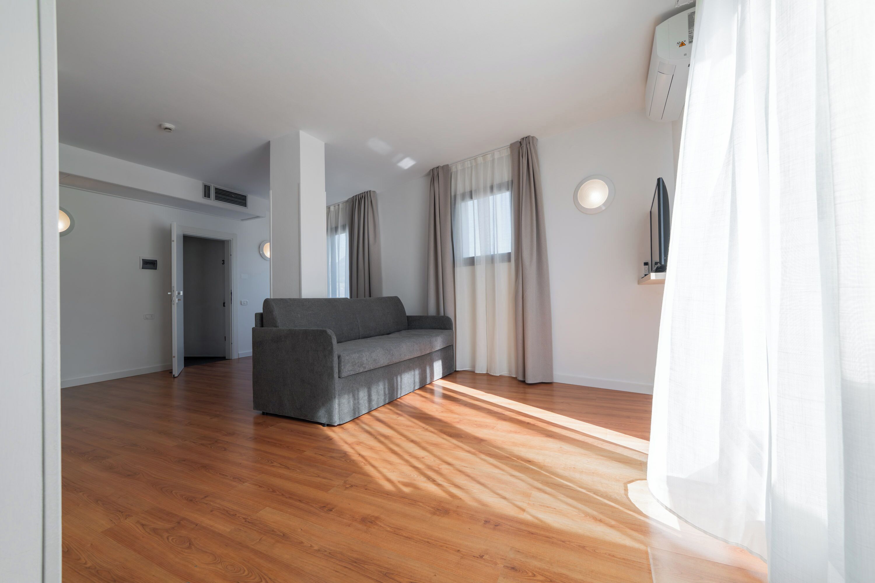 A modern living room with a grey sofa, wooden flooring, and a large window with curtains.