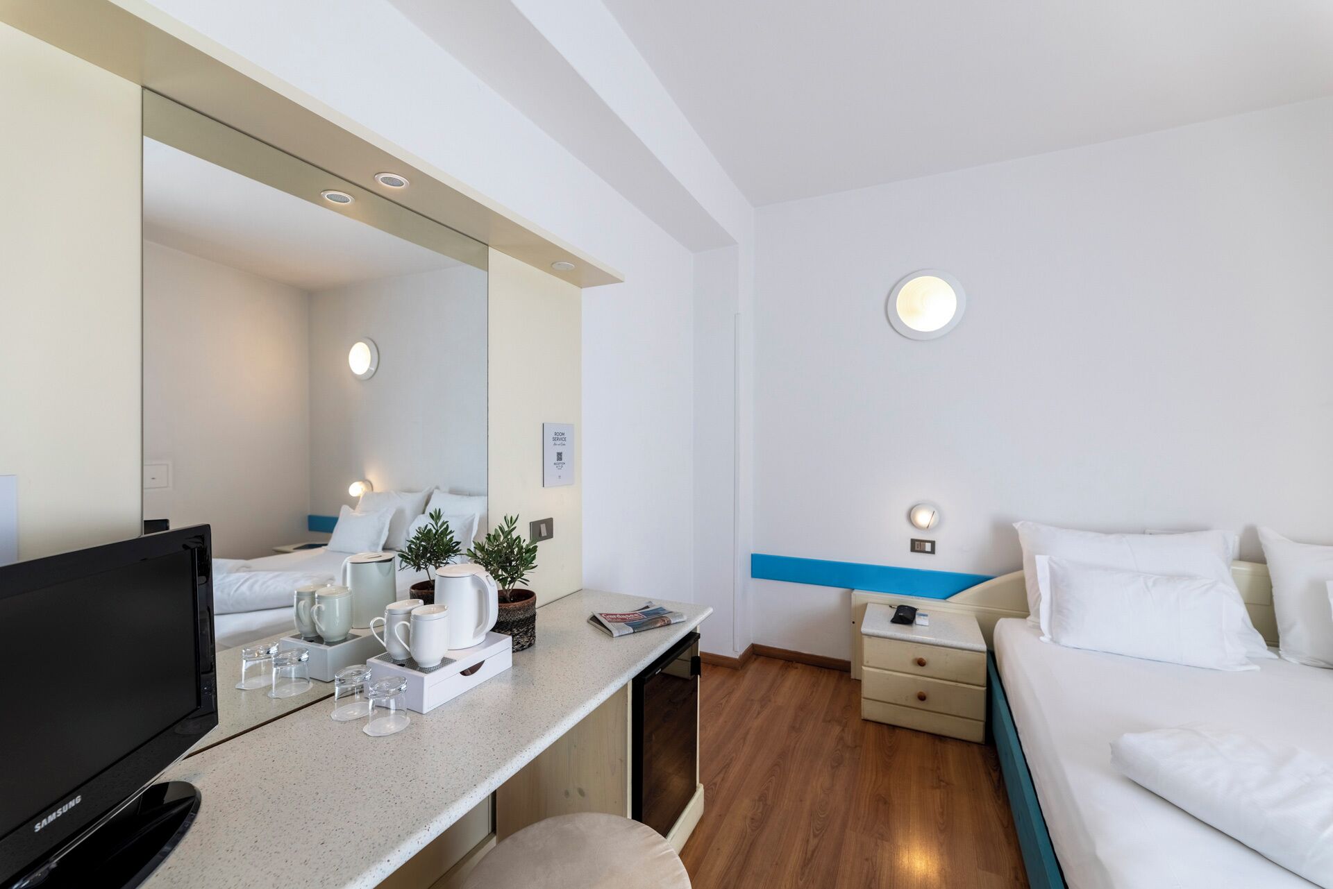 A modern hotel room with a flat-screen TV, a coffee station, a bed, and a bedside table with a lamp.