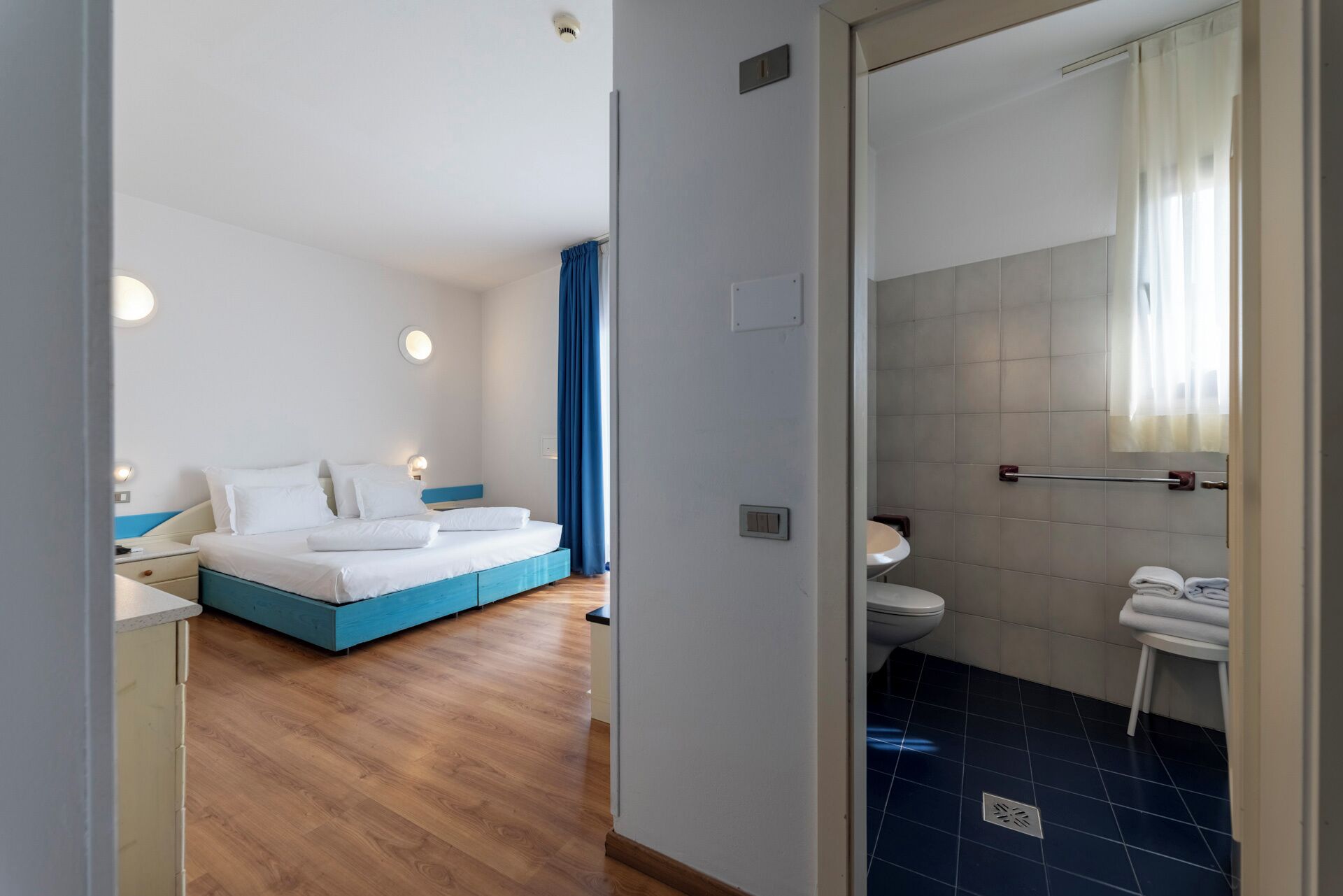 Una moderna camera d'albergo con pavimento in legno, un letto con testiera blu, un bagno bianco con doccia rivestita in piastrelle blu e un lavandino bianco.