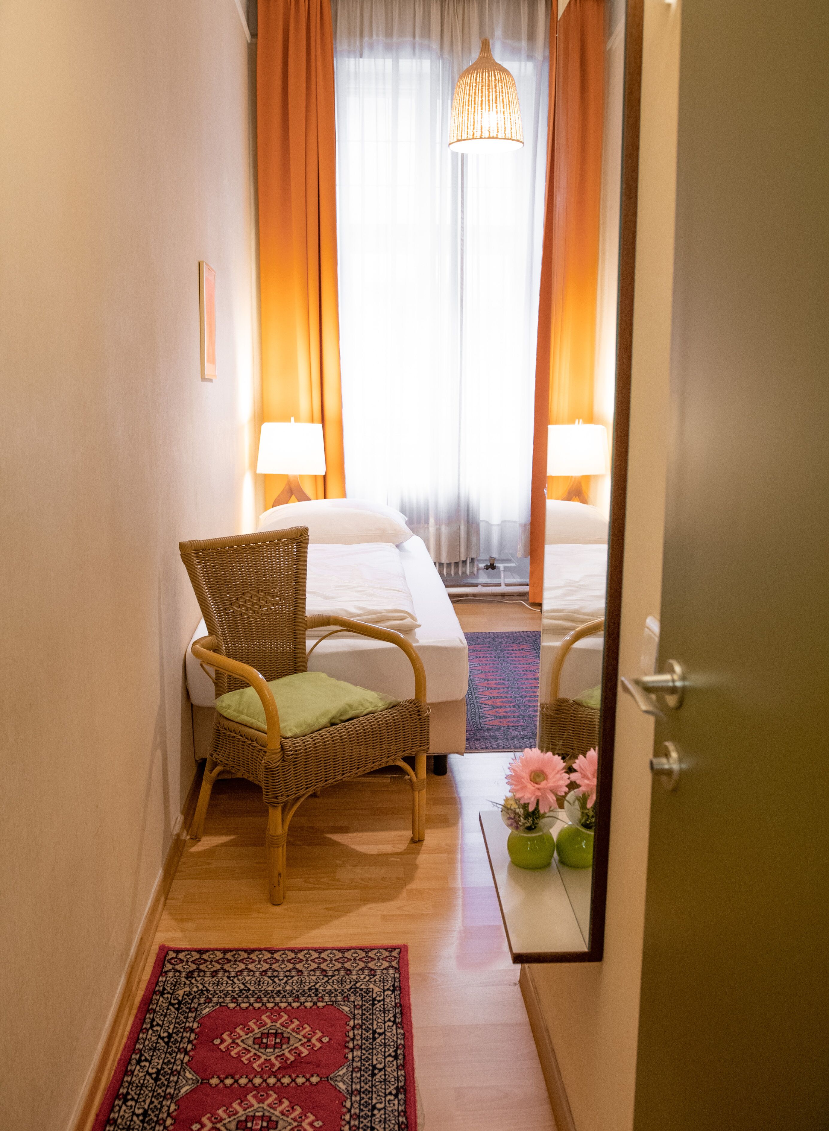Ett hotellrum med en flätad fåtölj, en säng, ett sängbord, en spegel och ett fönster med gardiner.