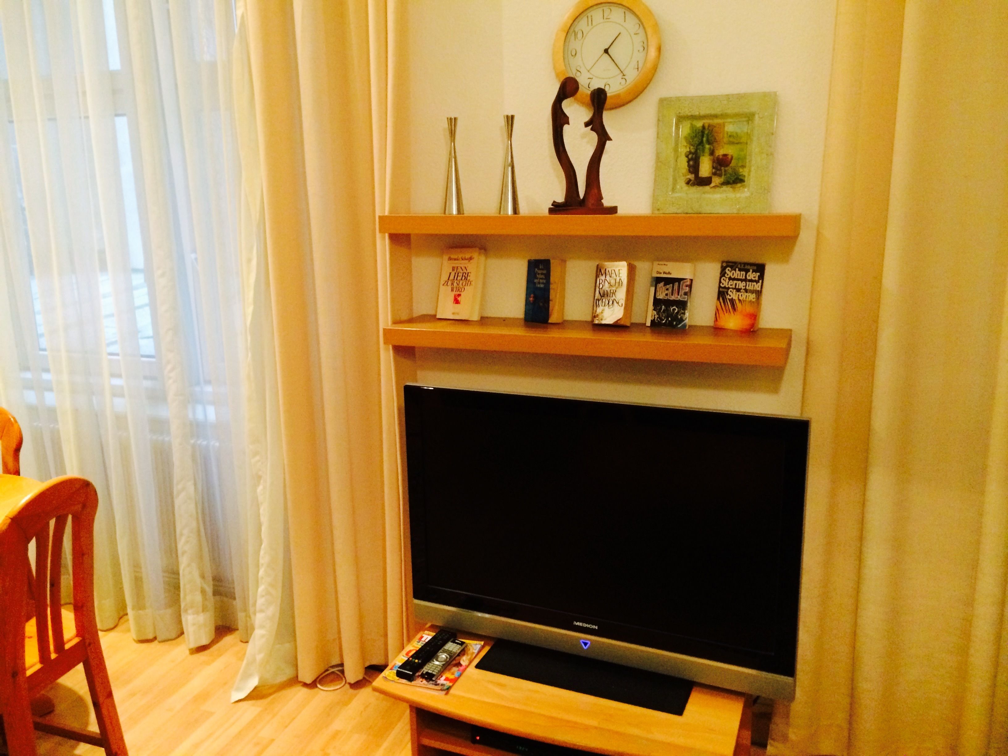 Una sala de estar con una televisión de pantalla plana, un estante de madera con libros, un reloj y un cuadro enmarcado.