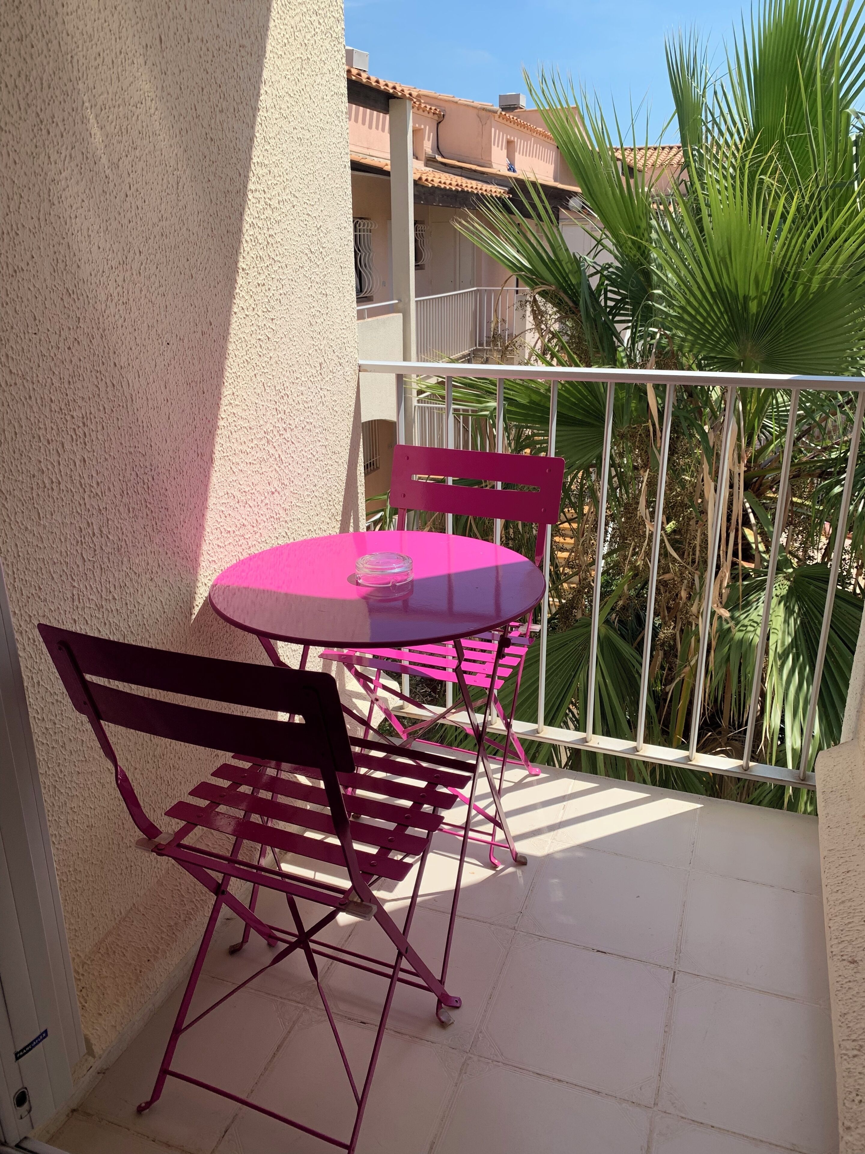 Un petit balcon avec une table ronde et deux chaises, un pot de fleurs et une vue sur un immeuble et des palmiers.
