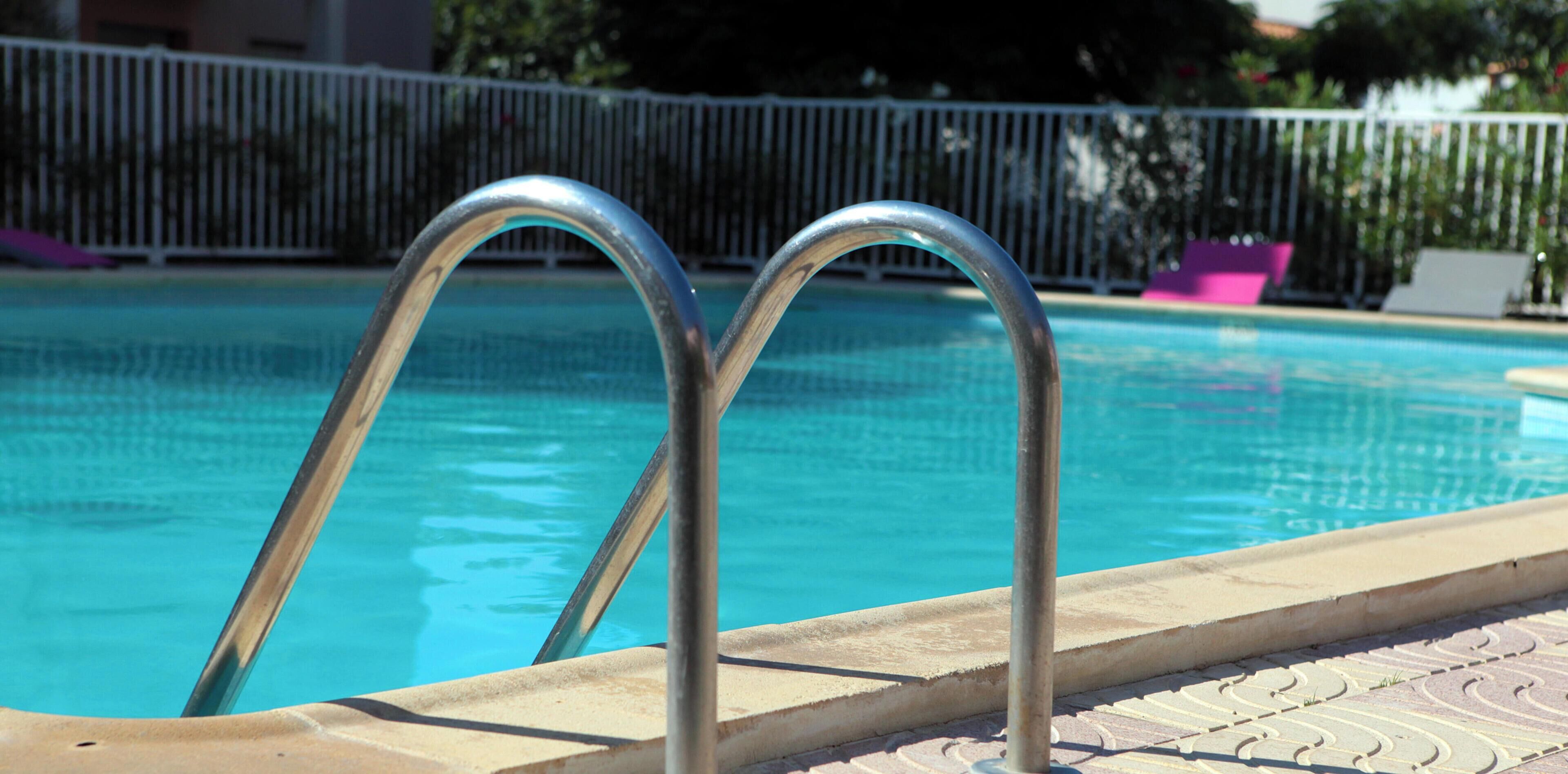 Una piscina con barandillas de metal, un suelo de baldosas y una valla blanca al fondo.