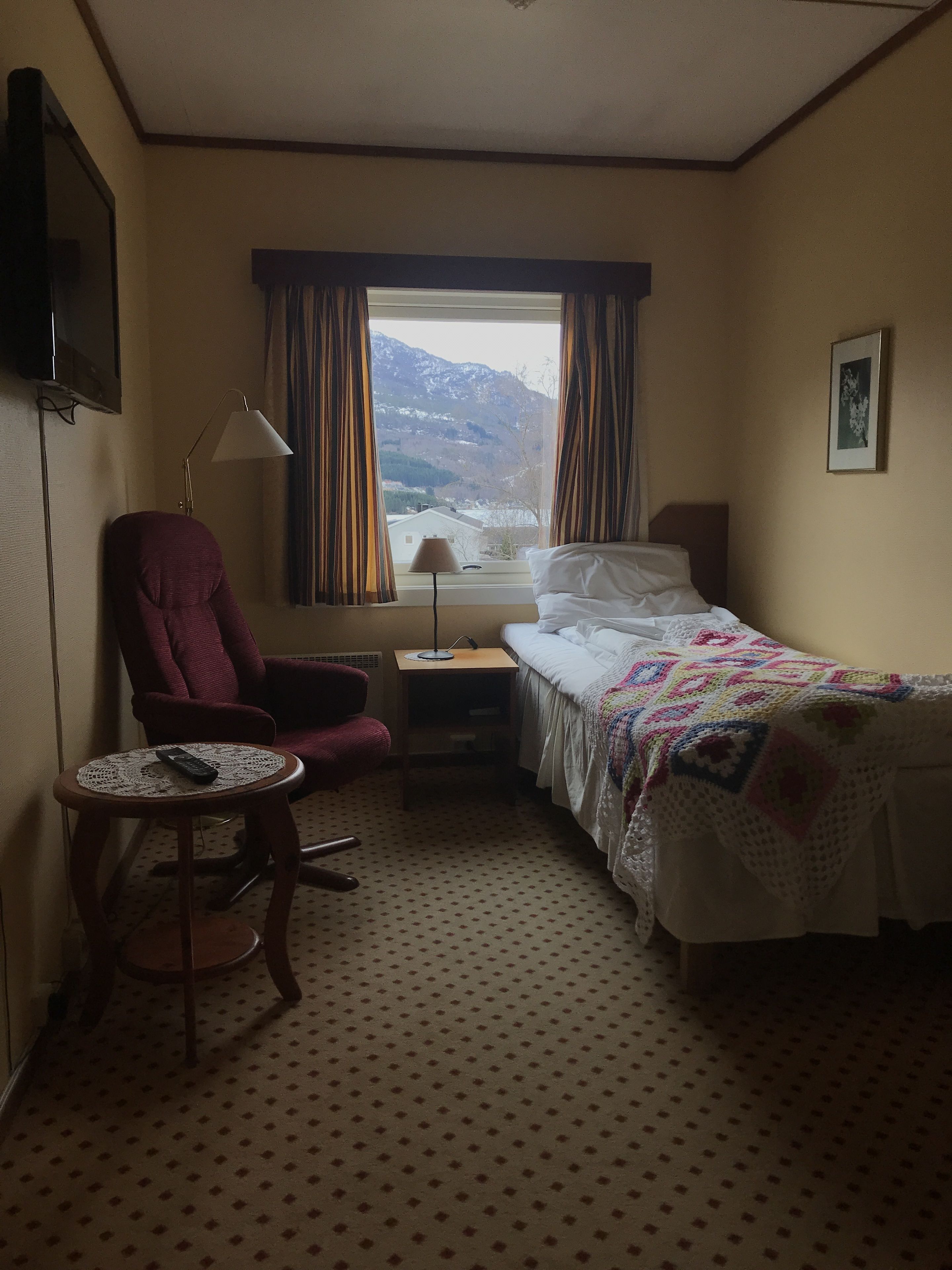 A hotel room with a bed, a chair, a small table, a nightstand, a lamp, a window with curtains, and a picture on the wall.