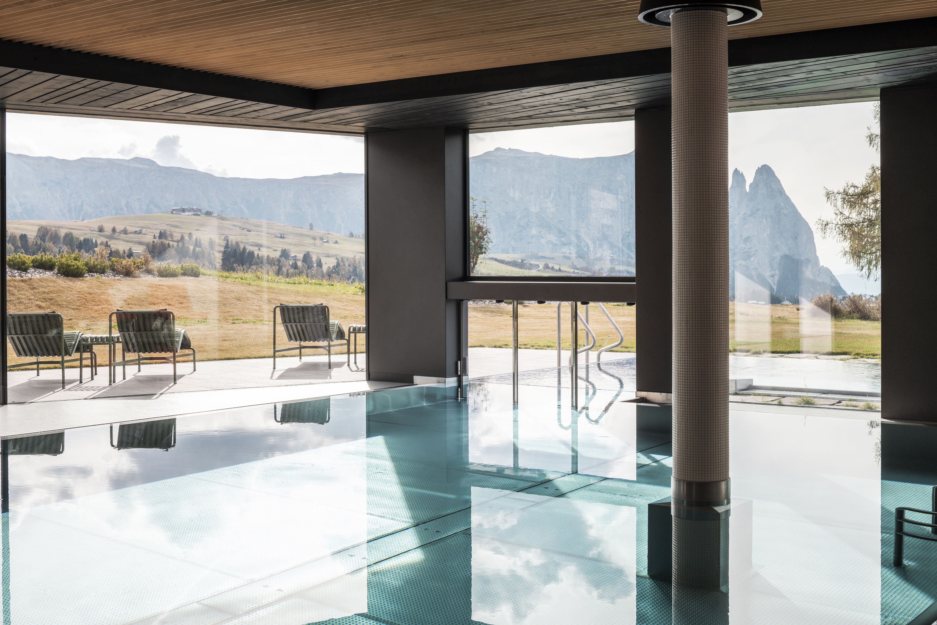 A modern indoor swimming pool with clear glass walls, a wooden ceiling, and a view of mountains.