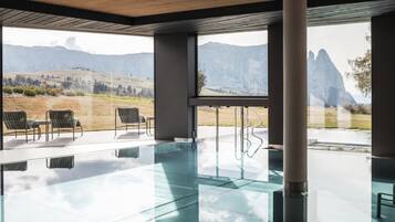 A modern indoor swimming pool with clear glass walls, a wooden ceiling, and a view of mountains.