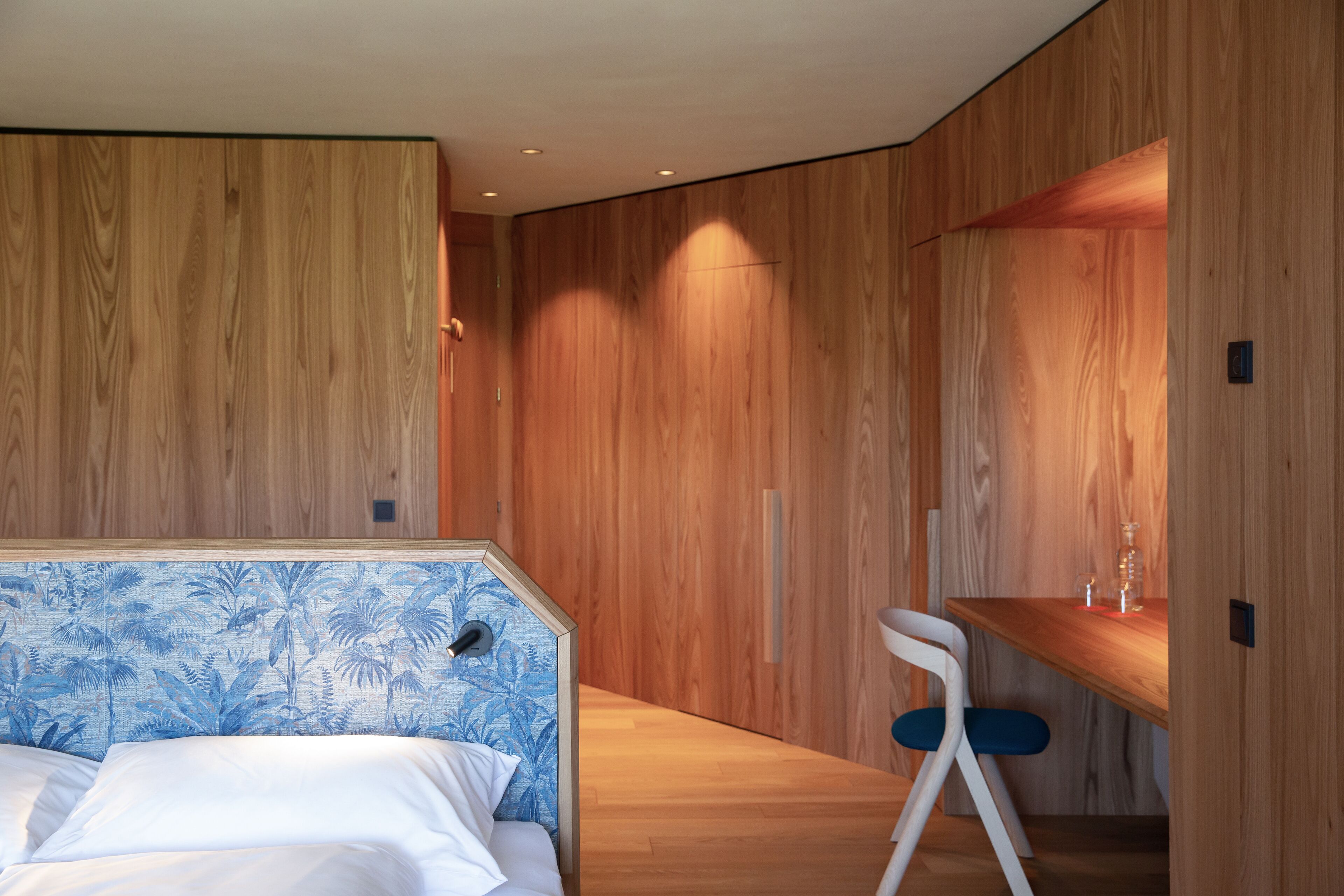 Ein modernes Schlafzimmer mit Holzwand, einem Bett mit blauem, floralem Kopfteil und einem kleinen Holztisch mit Stuhl.