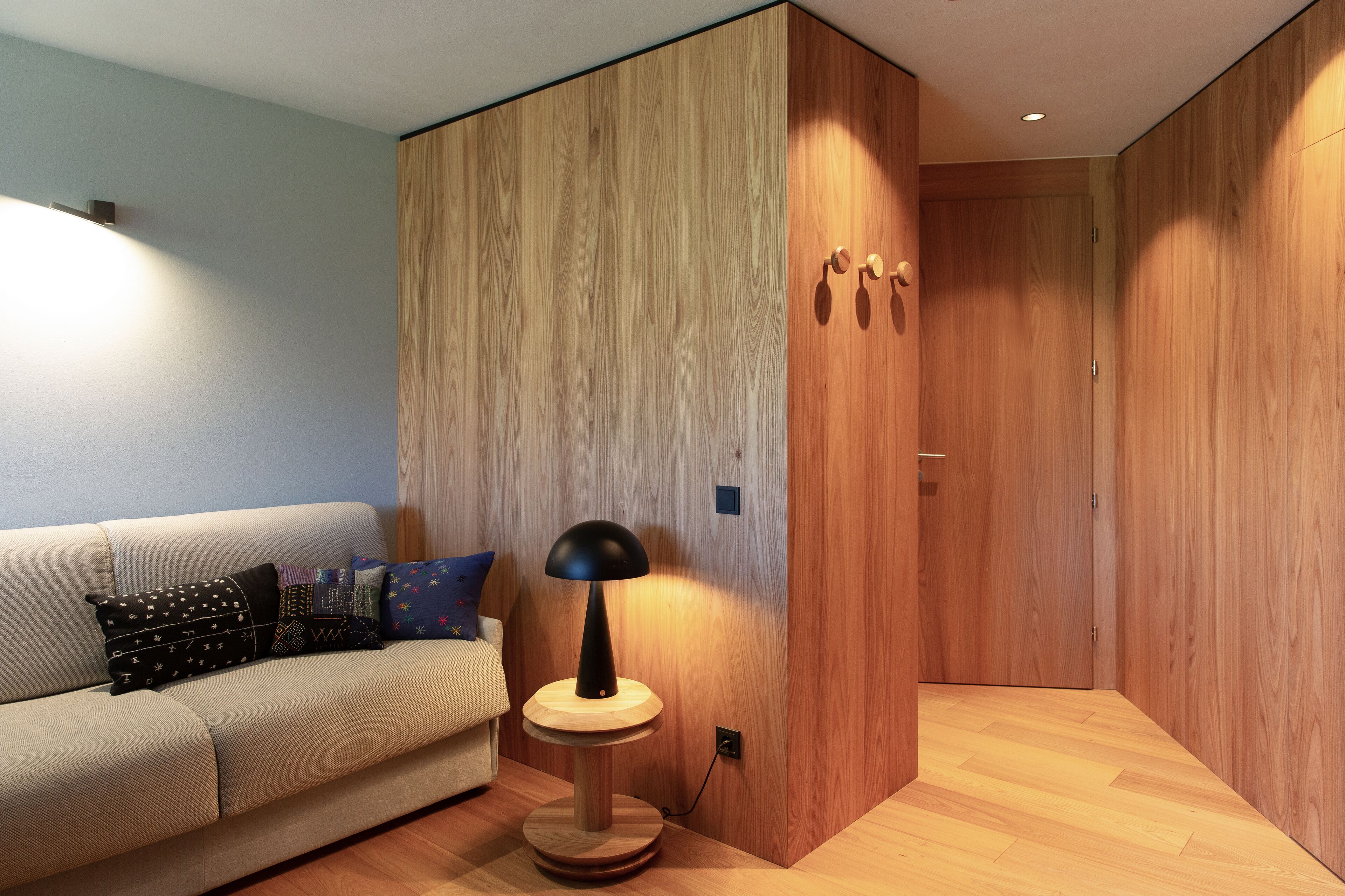 Ein modernes Zimmer mit Holzboden, einem runden Holztisch mit Lampe, einem beigen Sofa mit gemusterten Kissen und einer Holzwand.