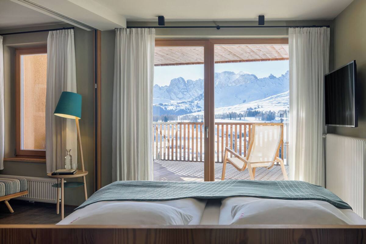 A bedroom with a bed, a bedside table with a lamp, a window with curtains, and a view of mountains through a balcony.