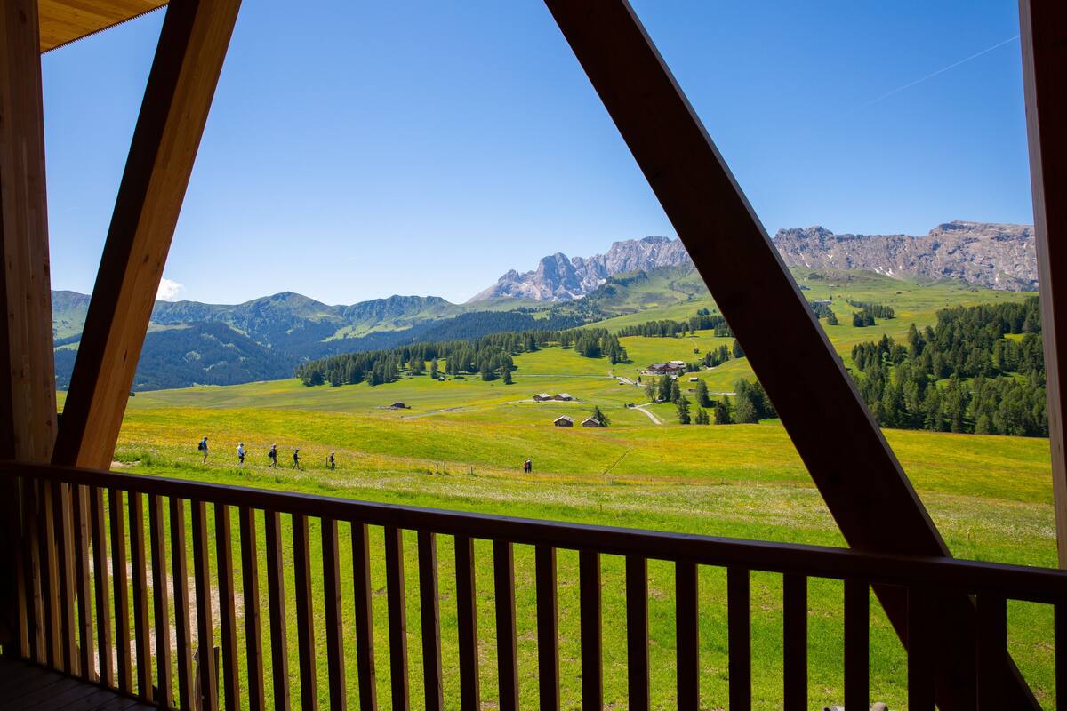 A view from a wooden balcony overlooking a green valley with houses and mountains in the background.