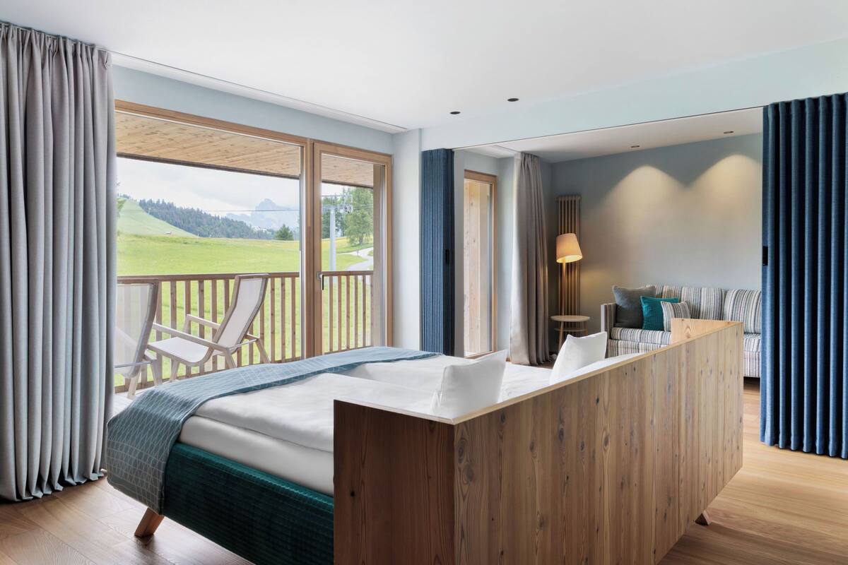 A bedroom with a large bed, a wooden desk, a balcony with a chair, and a view of a green landscape.