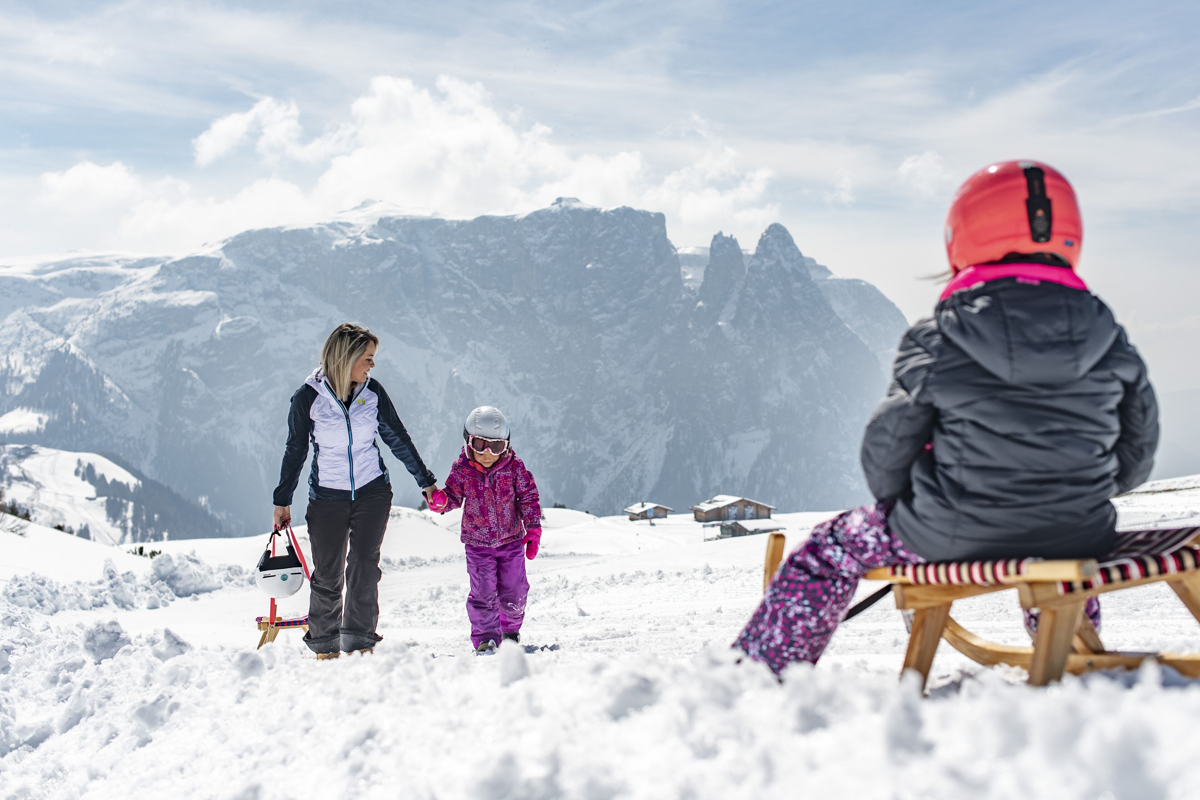 A snowy mountain scene with a person in a black jacket and red helmet sitting on a sled, another person in a purple outfit walking, and a third person in a white jacket holding a child's hand.
