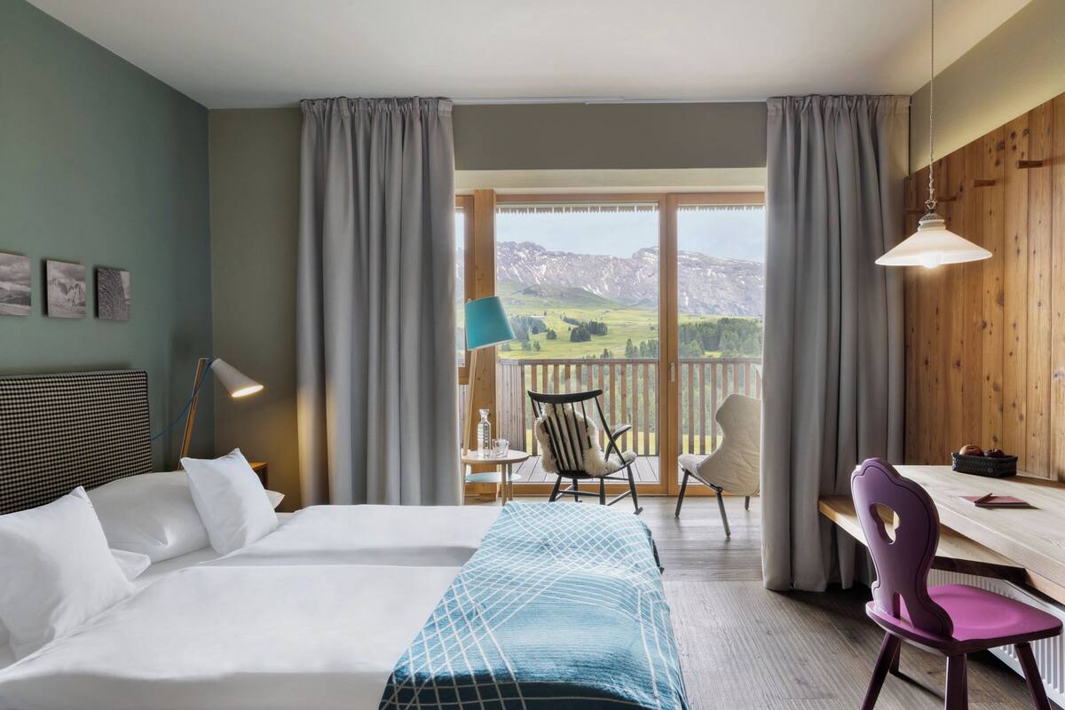A hotel room with a large bed, a desk, a chair, and a view of mountains through the window.